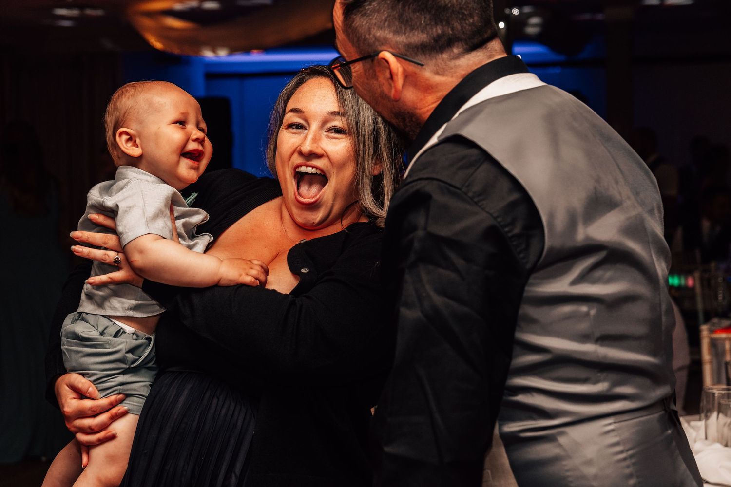 Joyful woman holds laughing baby while man in gray suit leans in at wedding reception.