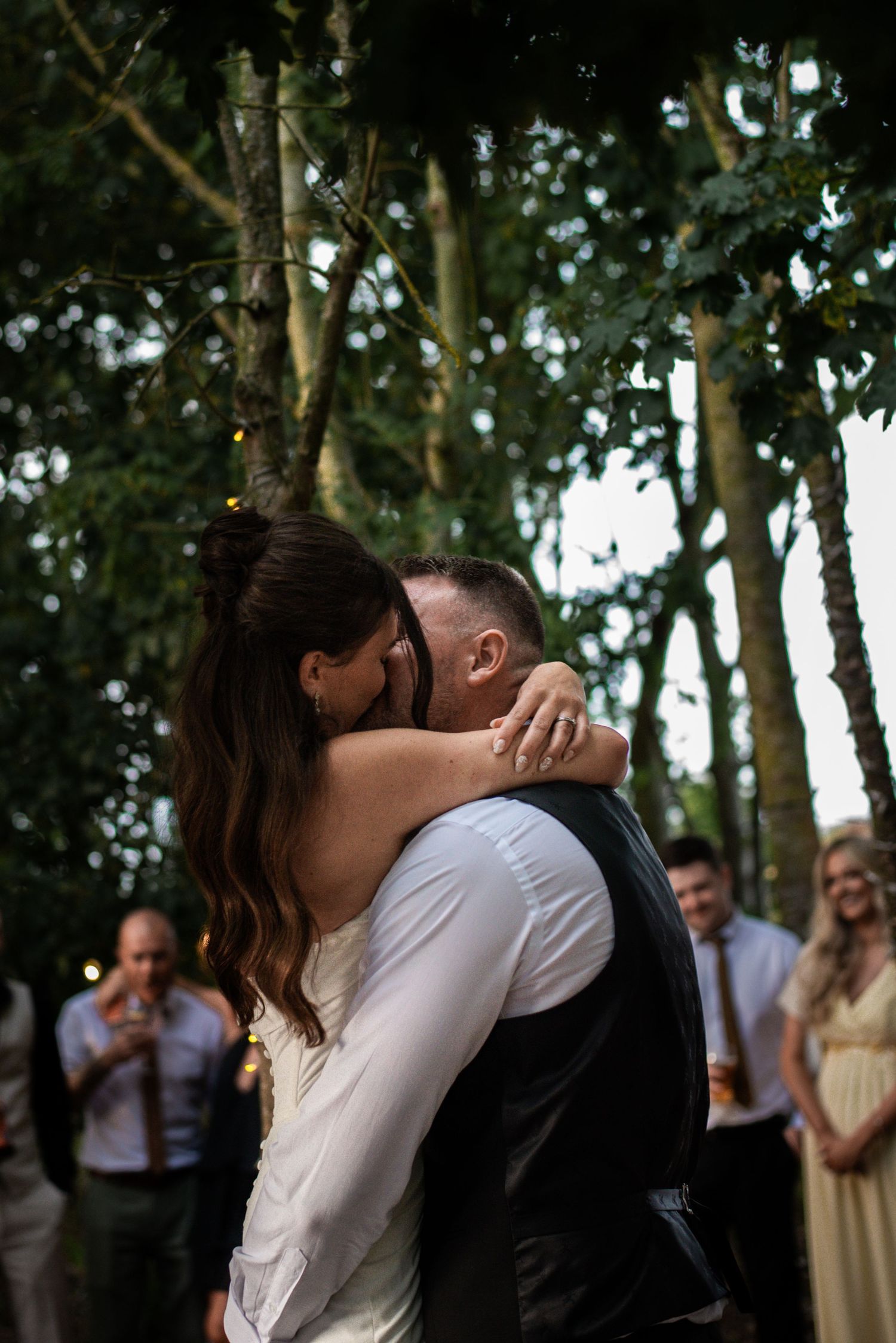Bride and groom share a romantic first dance embrace outdoors surrounded by trees and wedding guests.