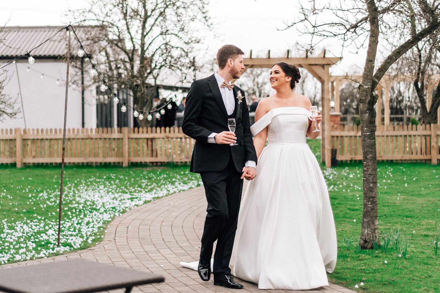 Bride and groom walking together outdoors, smiling, wearing wedding attire near a garden with white petals.