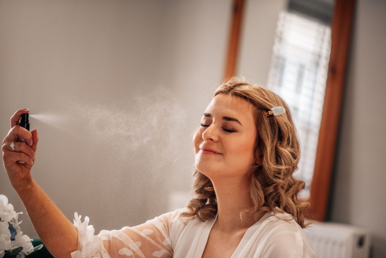 Smiling bride in white robe spraying perfume with eyes closed during wedding morning preparation.