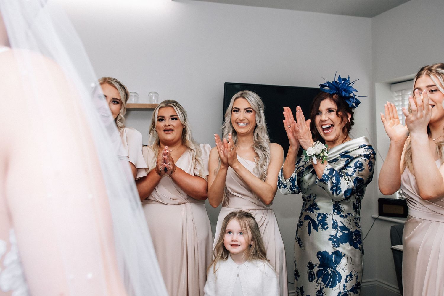Bridesmaids in champagne dresses and woman in floral hat clapping joyfully as bride reveals wedding dress.
