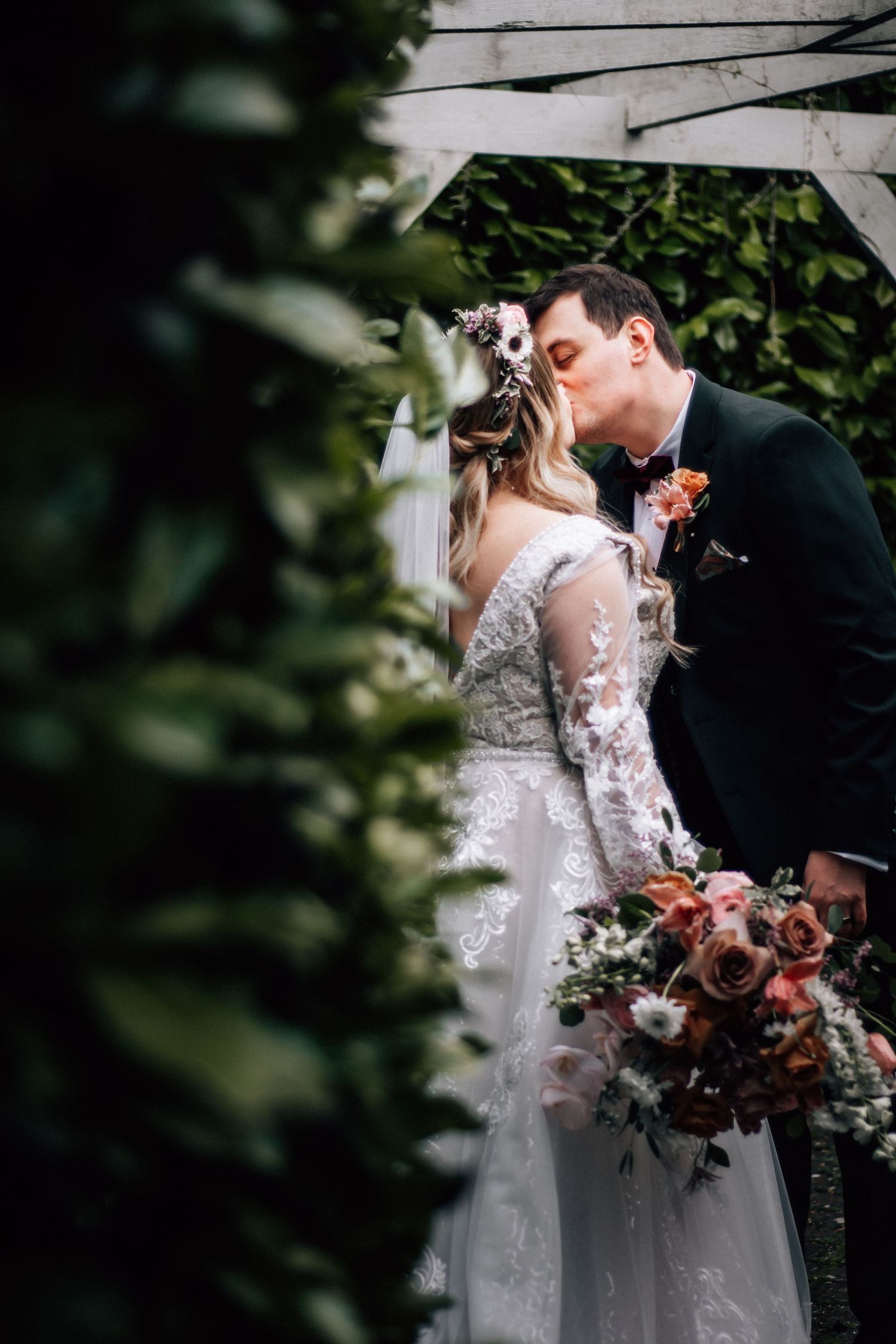 Bride and groom sharing a kiss under a white arbor surrounded by lush greenery on their wedding day.