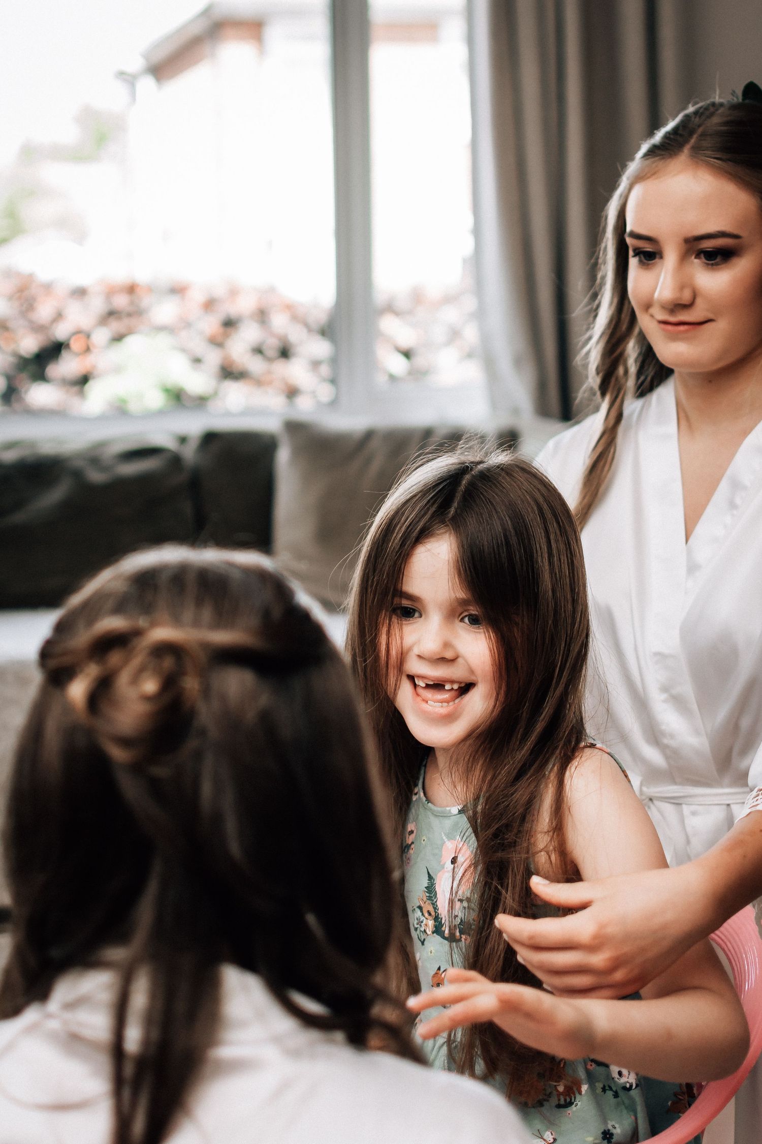 Young girl smiling with missing teeth as woman in white robe braids her hair indoors near window.