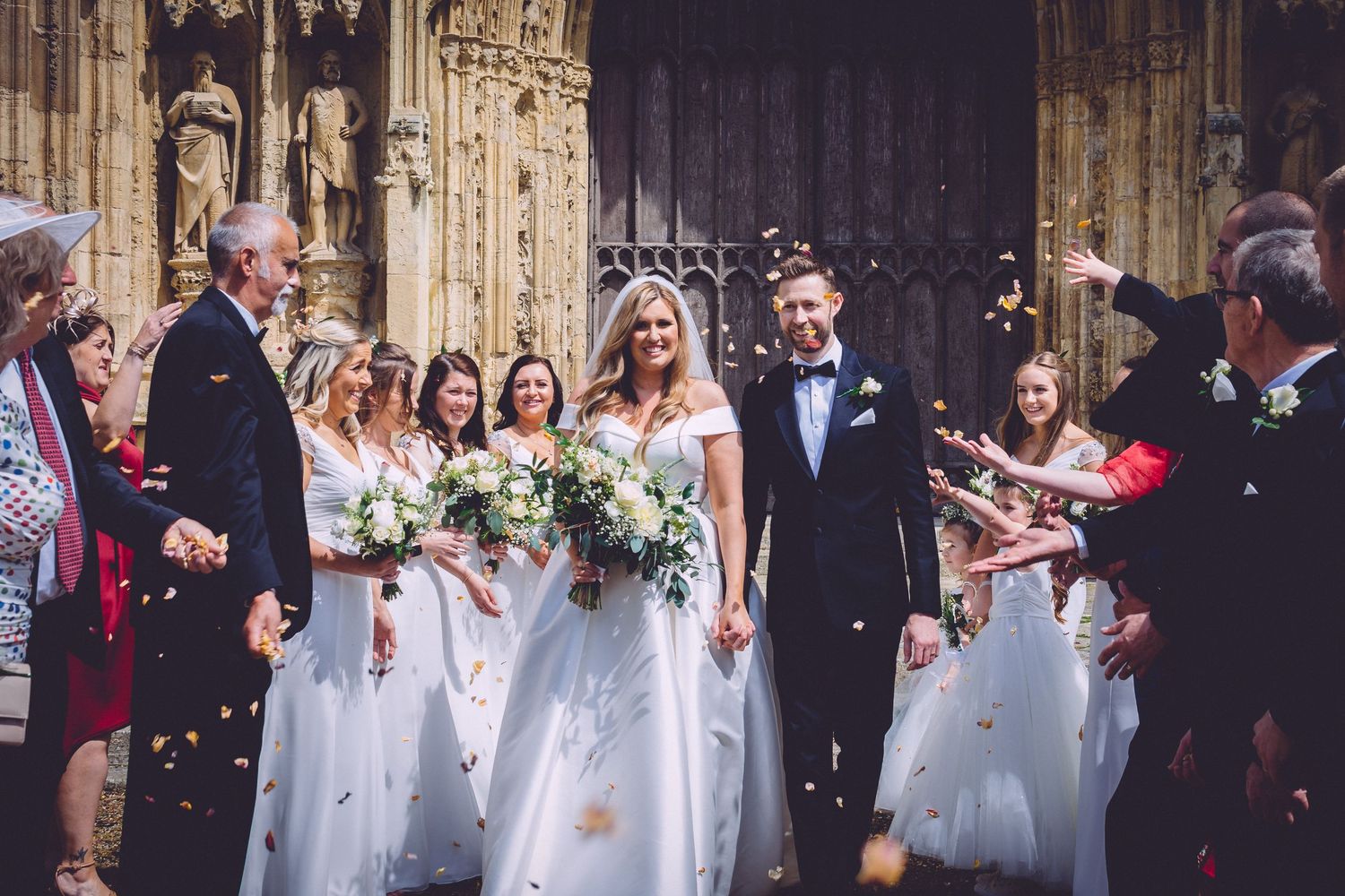 Bride and groom exit church surrounded by wedding party as guests throw confetti outdoors.