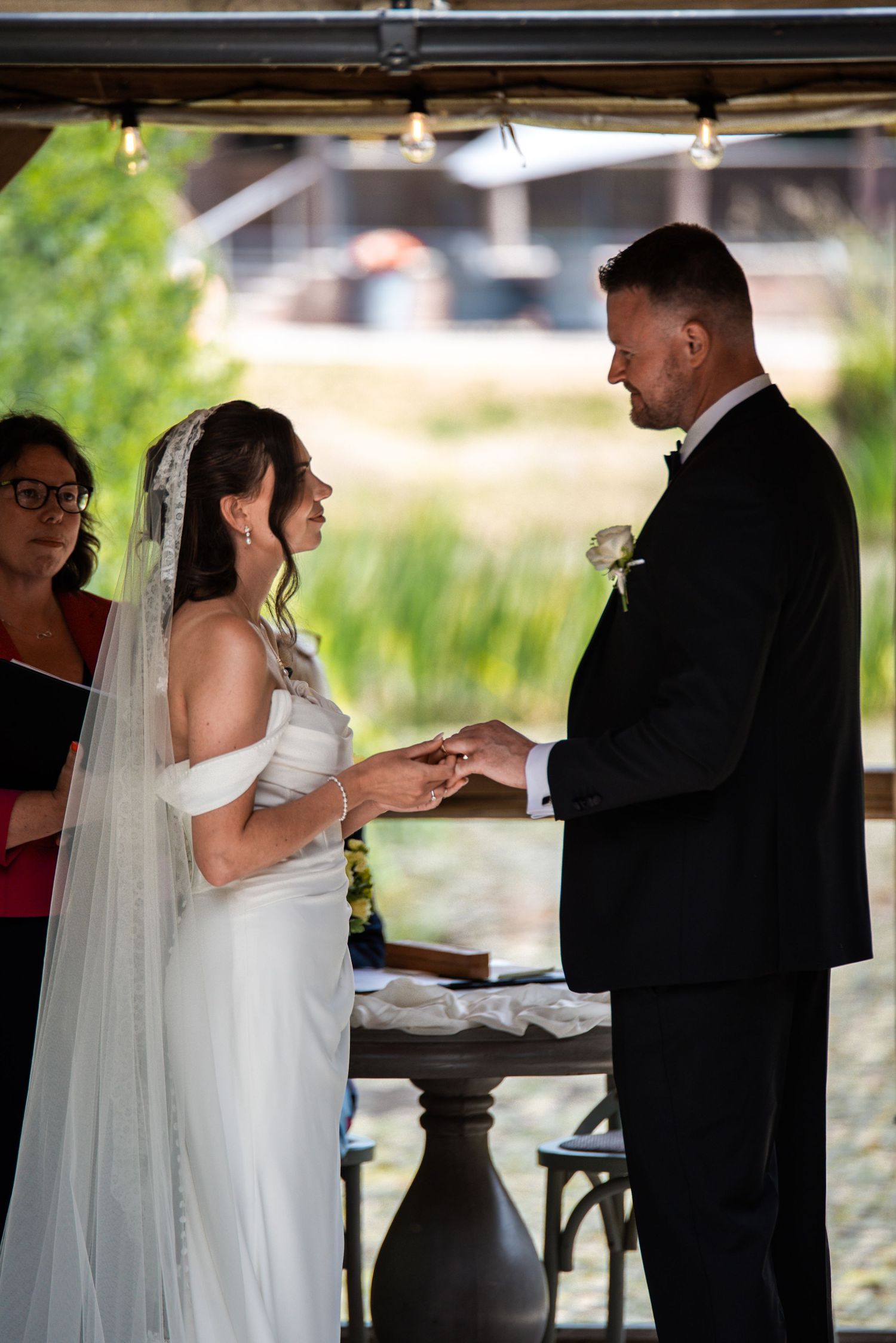 Bride and groom exchange rings during outdoor wedding ceremony, officiant visible in background near lush greenery.