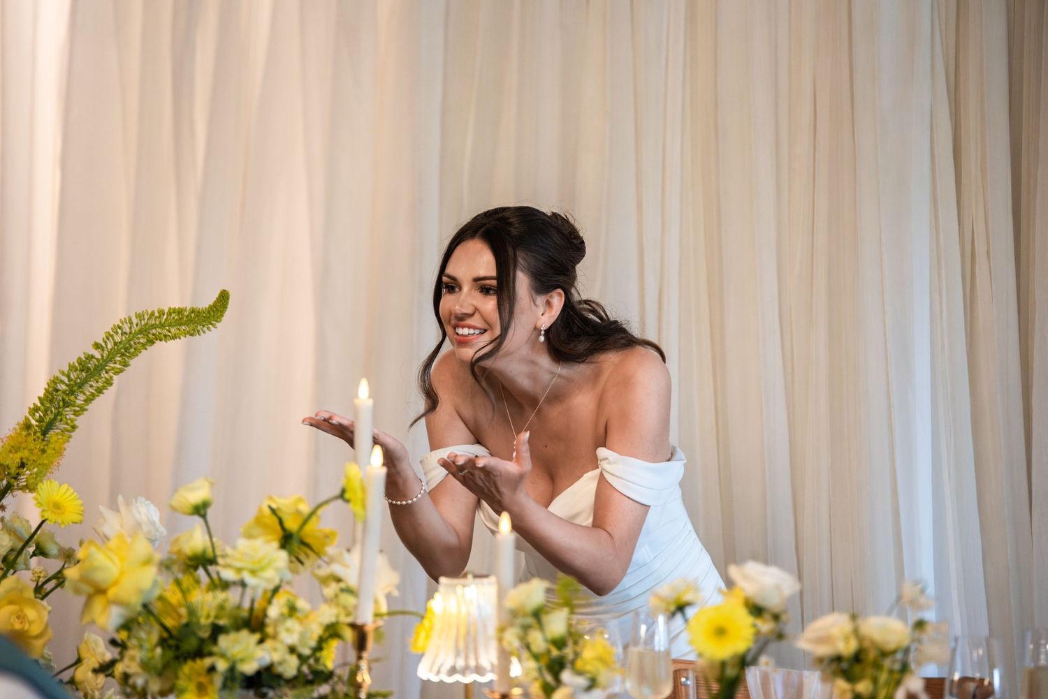 Bride in white off-shoulder gown laughing at candlelit table with yellow floral arrangements at elegant wedding reception.
