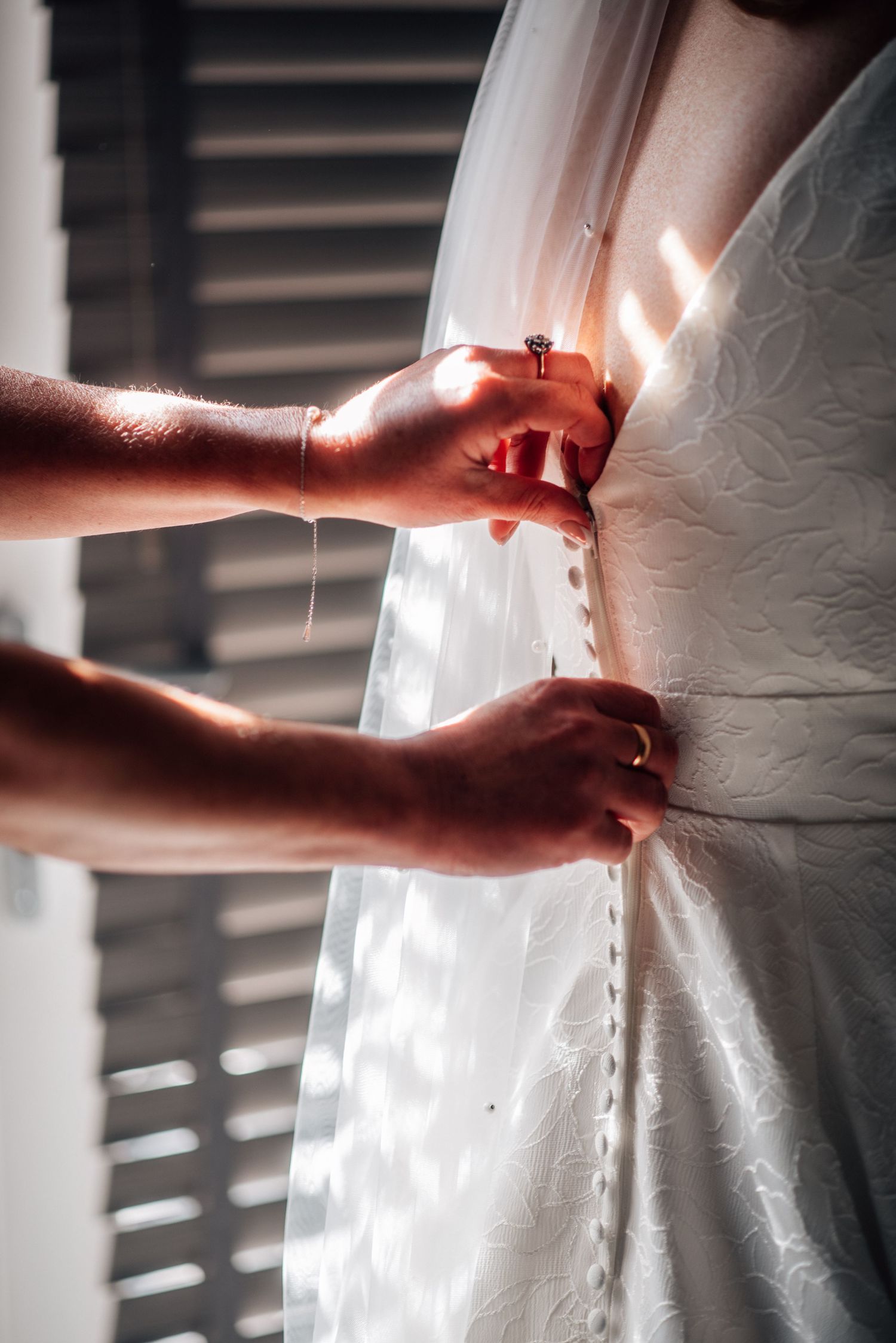 Hands fastening delicate buttons on a white lace wedding dress with veil in soft natural window light.