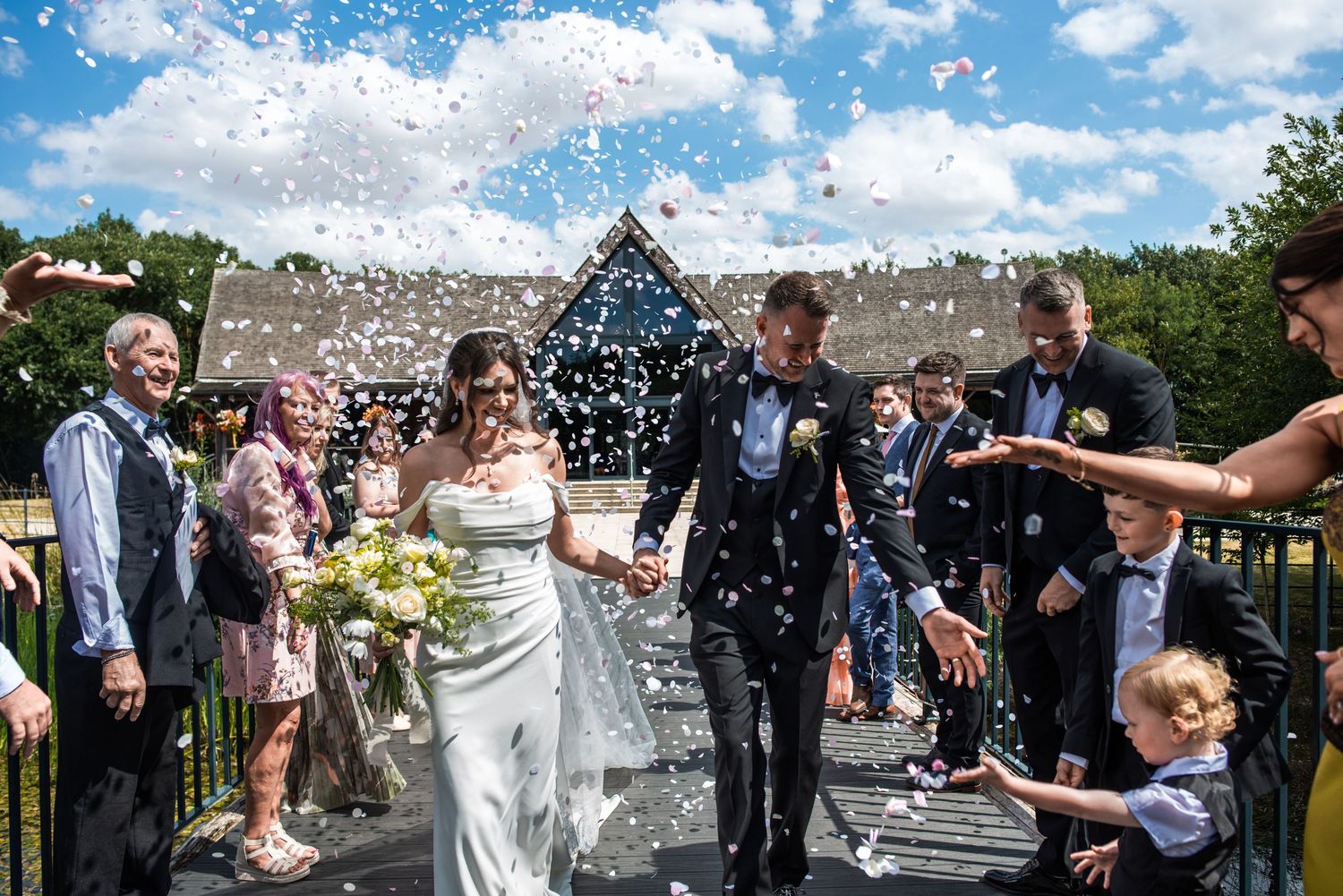 Bride and groom walk through confetti shower surrounded by smiling wedding guests outdoors on a sunny day.