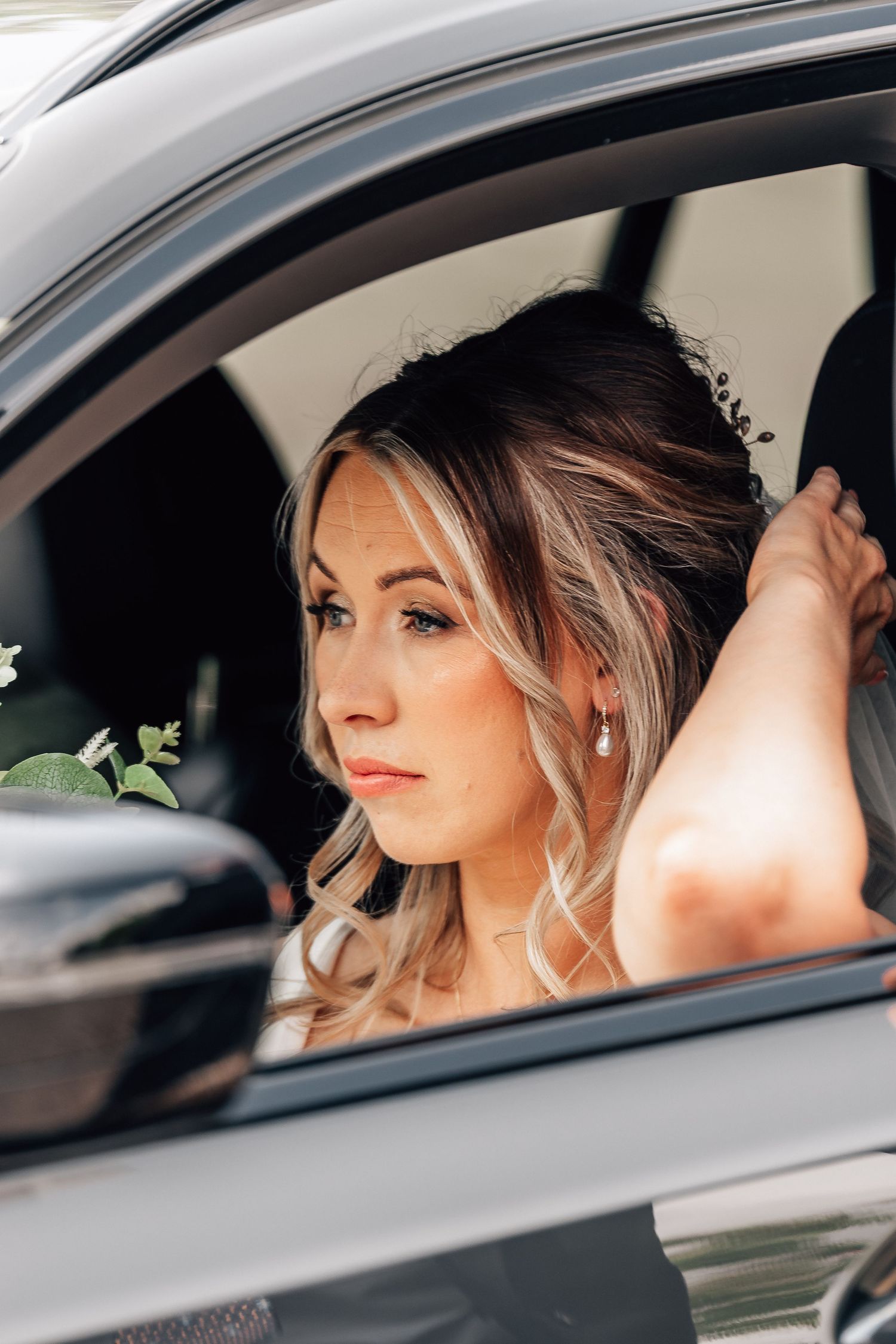 Elegant bride with blonde hair and pearl earrings gazing out of a car window on her wedding day.