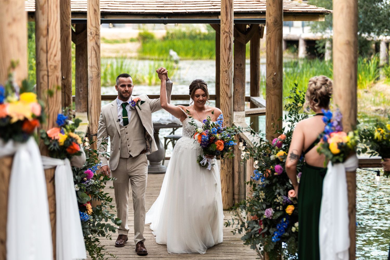 Joyful newlyweds celebrate on a decorated wooden dock, bride raising arms holding colorful bouquet, groom in tan suit.