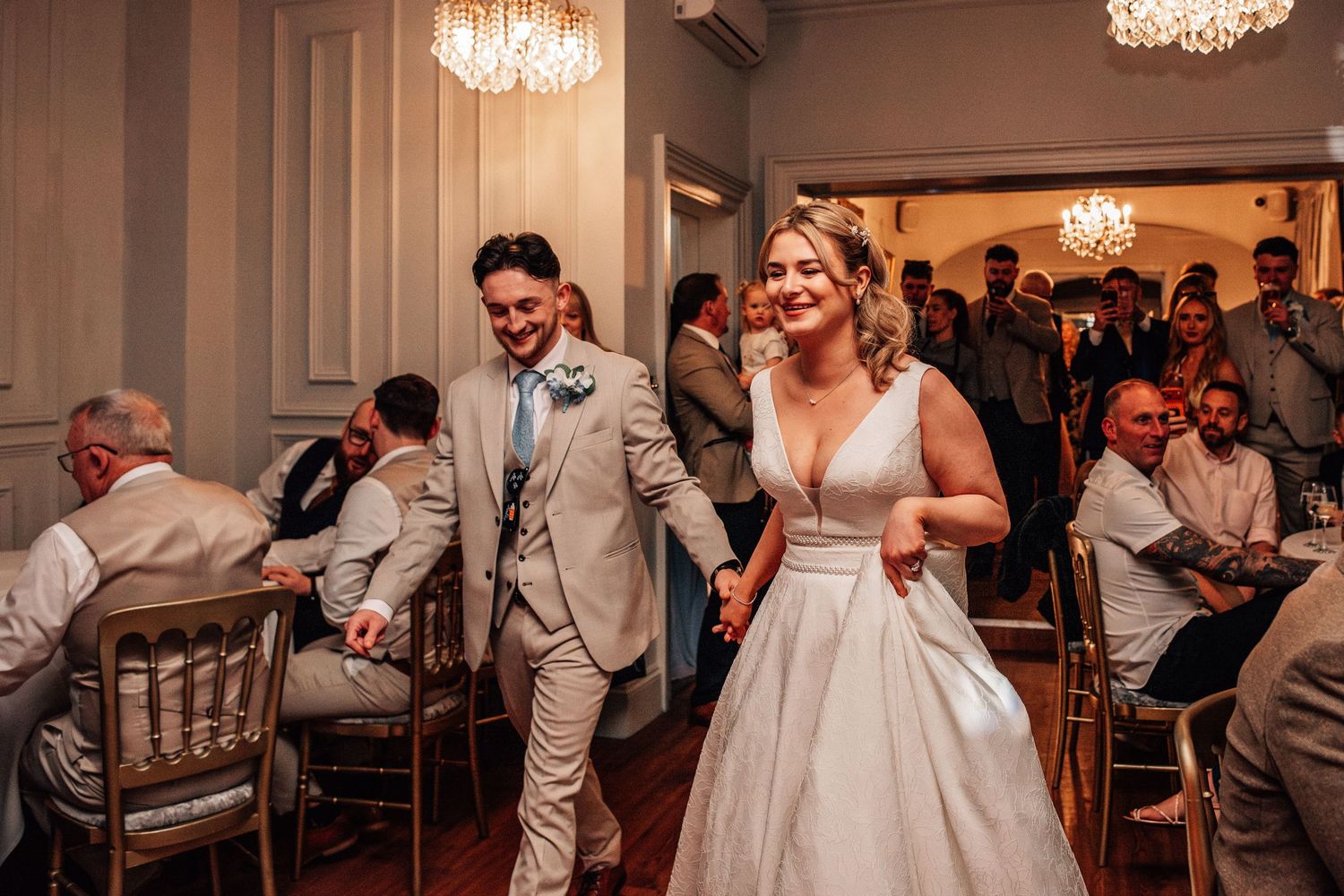 Bride and groom enter wedding reception hall, smiling and holding hands, guests seated at tables under crystal chandeliers.