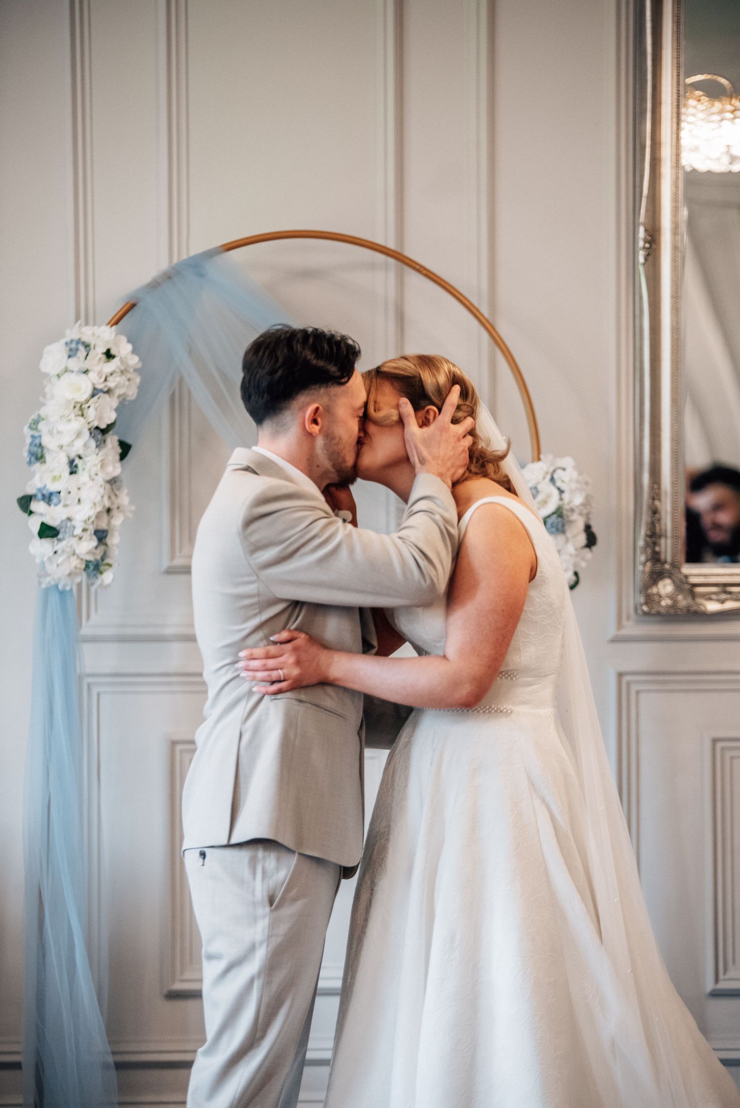 Bride and groom sharing first kiss under golden circular arch adorned with white flowers and blue tulle.