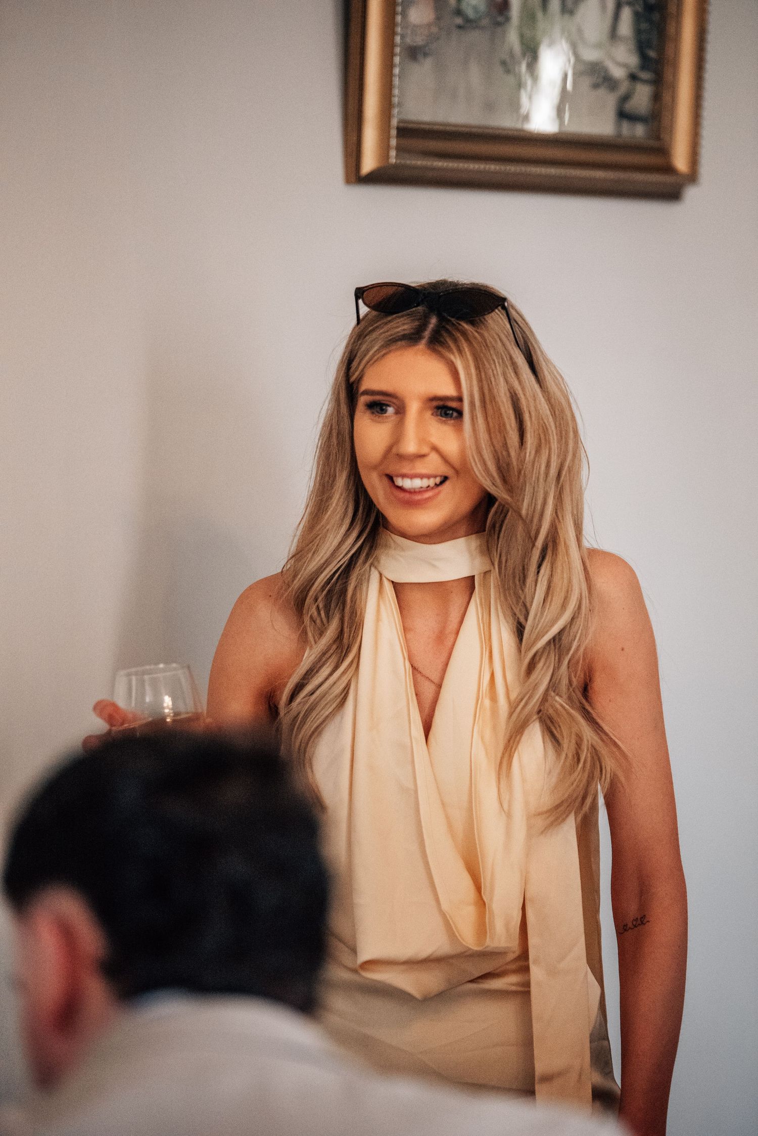 Smiling blonde woman with sunglasses on head holding wine glass wearing cream halter top indoors.