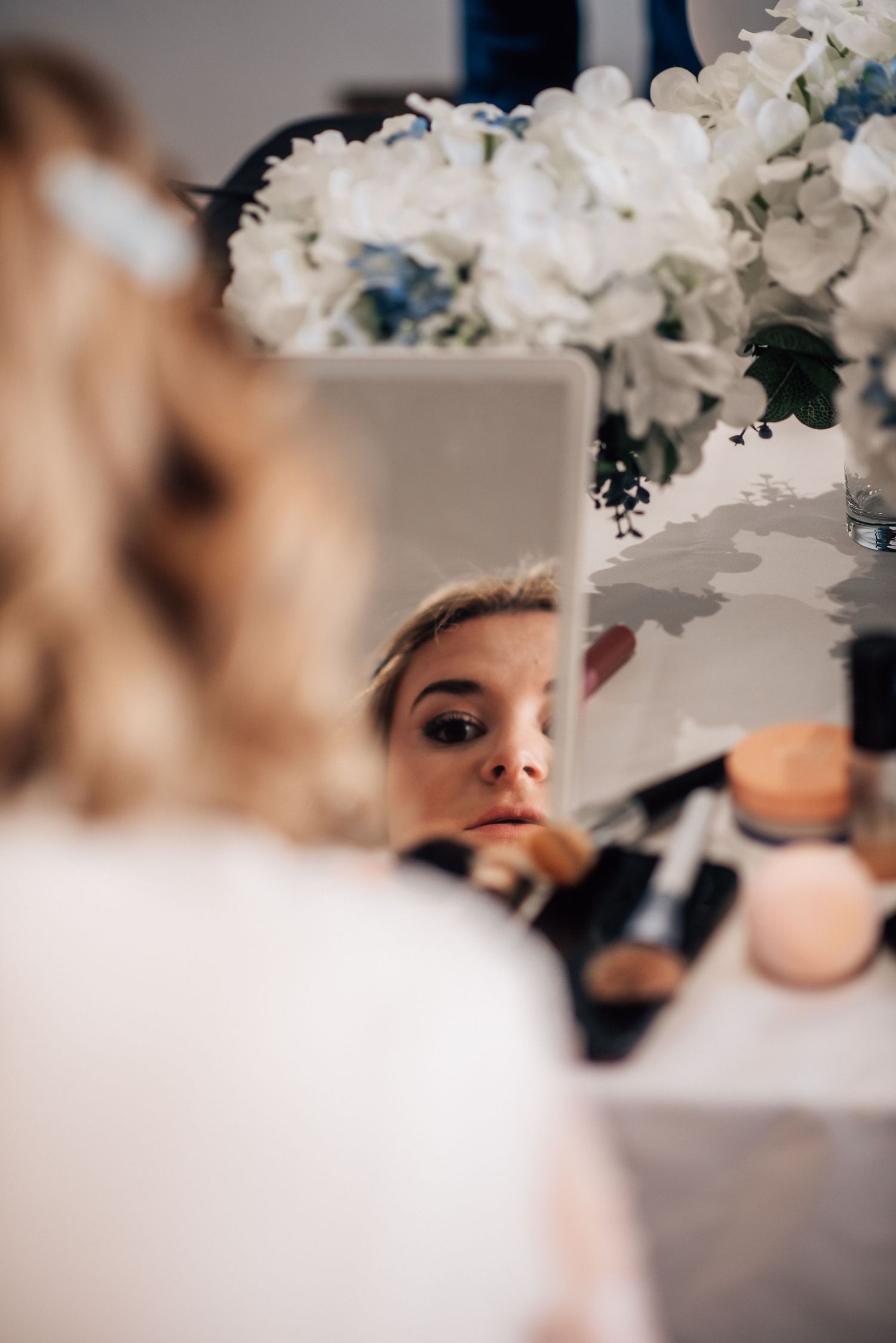 Bride getting makeup applied while checking her reflection in a mirror surrounded by white floral arrangements.