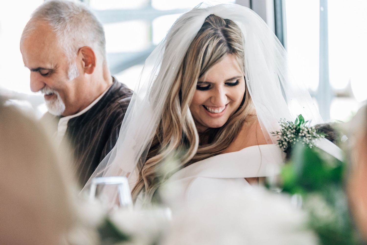 Smiling bride in white veil and off-shoulder gown seated at wedding reception table.
