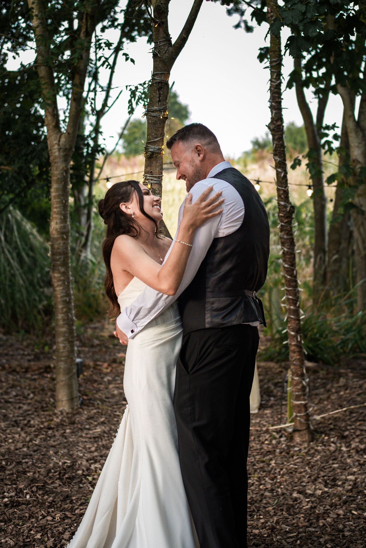 Bride and groom share a joyful embrace among trees with string lights at outdoor wedding.