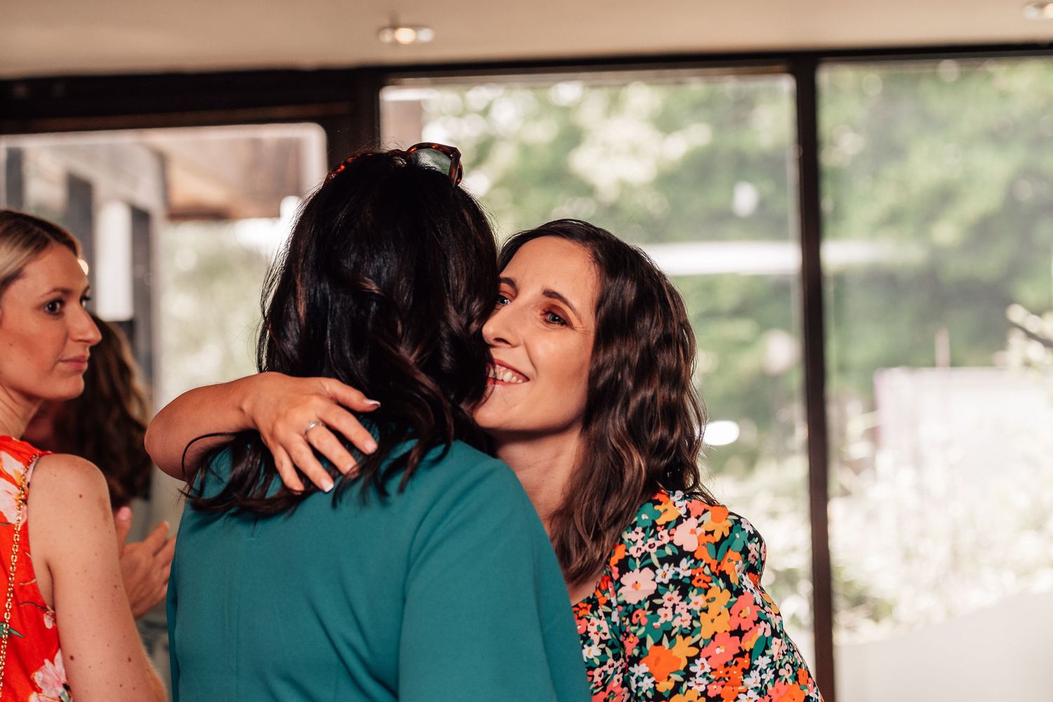 Woman in floral dress smiling while hugging someone at a social gathering indoors.