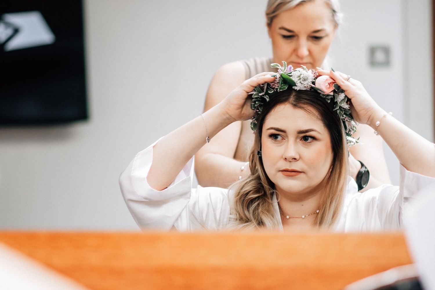 A bridesmaid places a floral crown on a bride's head as she sits in a white robe before her wedding.