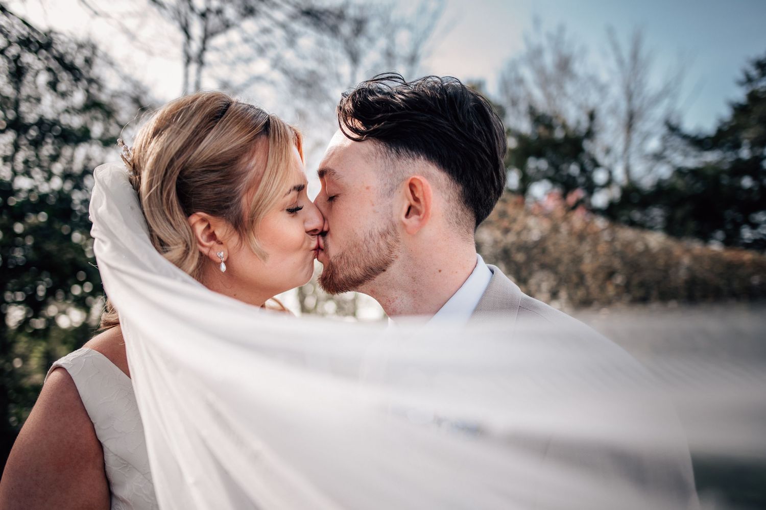 A bride and groom share a romantic kiss outdoors, with the wedding veil flowing in the breeze on their wedding day.
