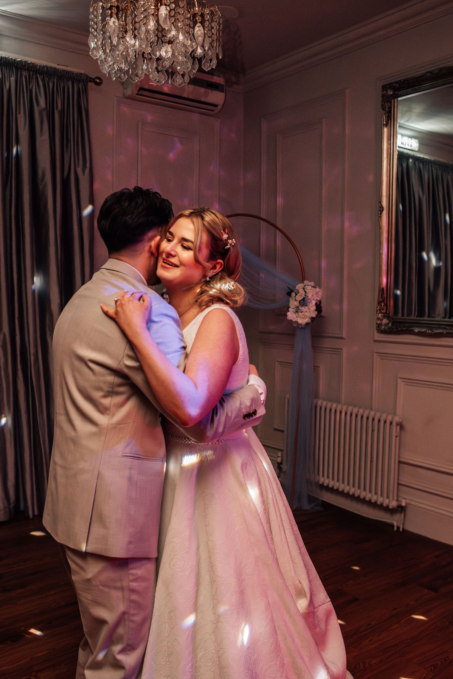 A bride and groom share their first dance in a softly lit elegant reception room with a crystal chandelier overhead.