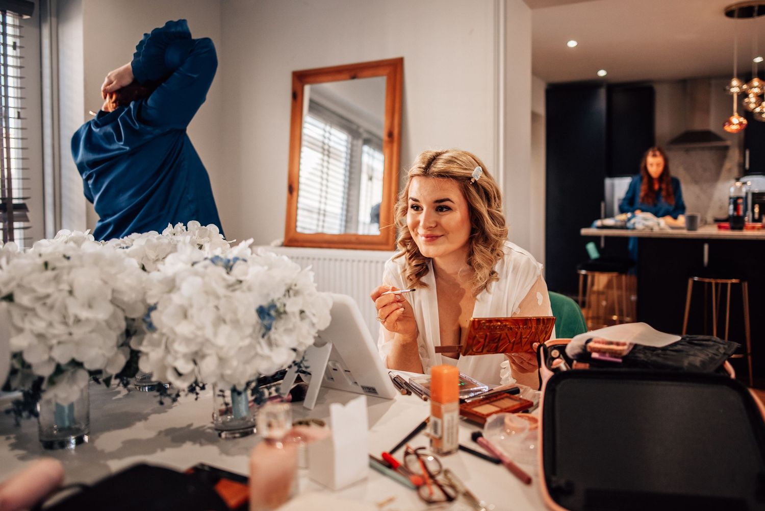 Bride applying makeup at a vanity mirror surrounded by white flowers and cosmetics, with bridesmaids preparing in background.