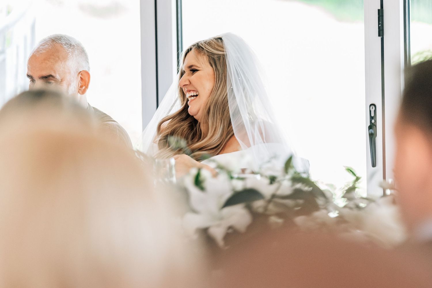 Smiling bride in white veil laughing joyfully at wedding reception table with floral arrangements.