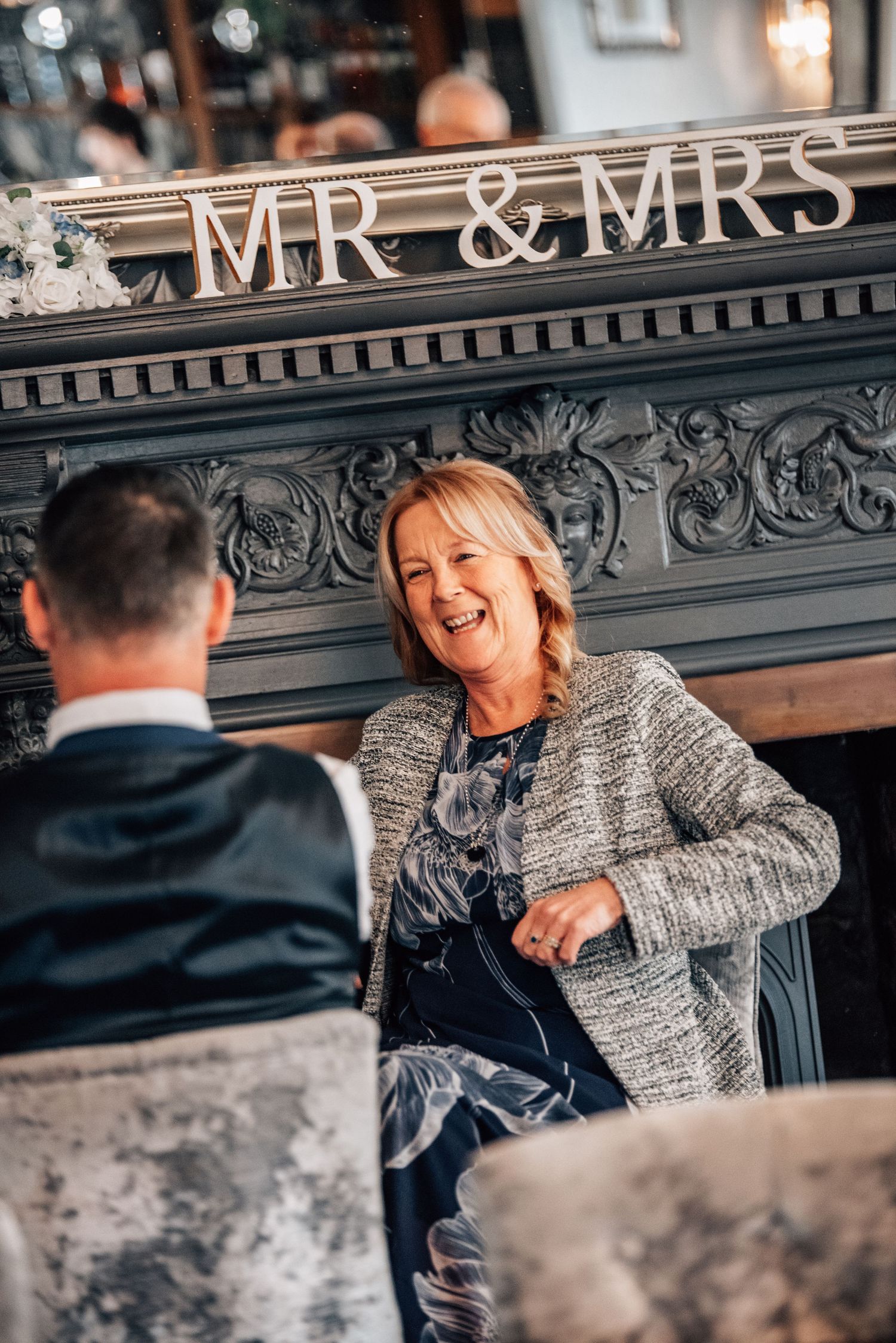 A smiling woman in a tweed jacket sits by an ornate fireplace with a Mr & Mrs sign at a wedding reception.