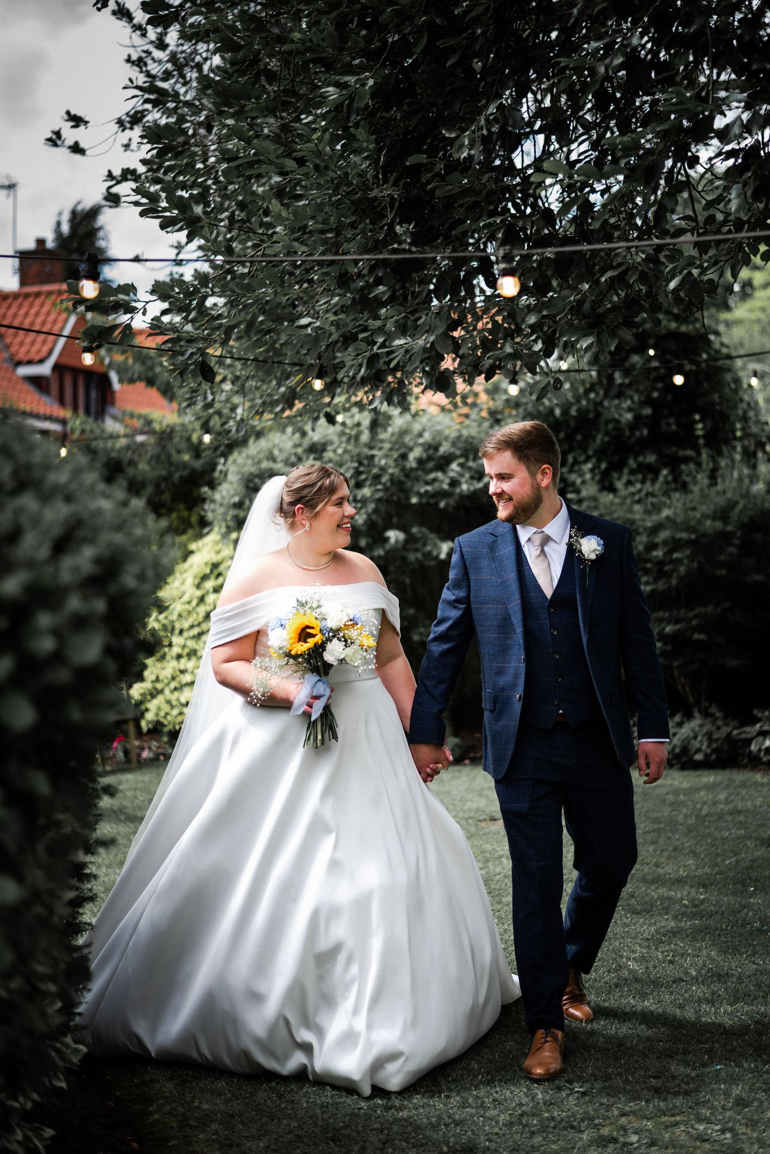Bride in white off-shoulder gown holds sunflower bouquet while walking hand-in-hand with groom in navy suit in garden.