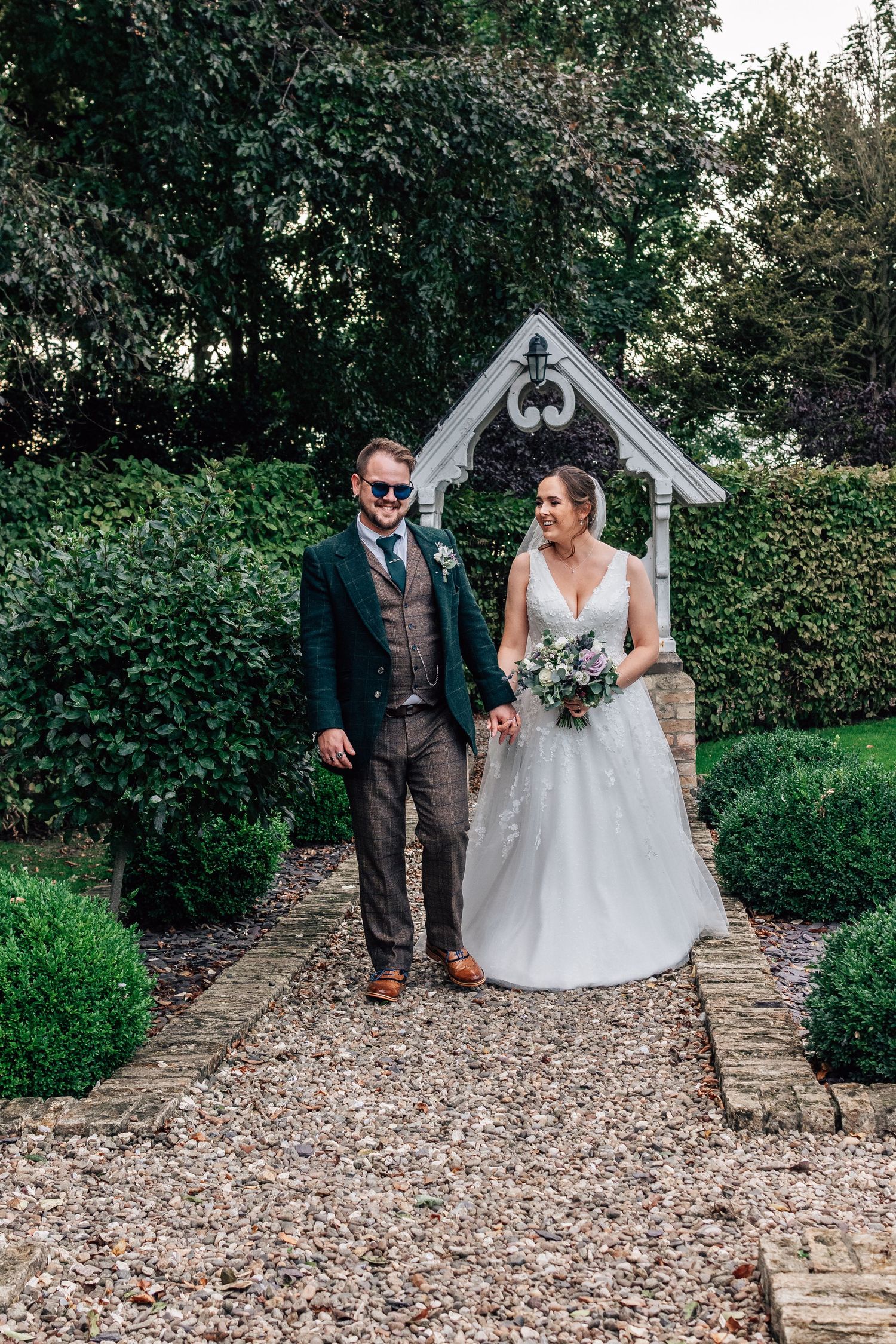 Bride and groom walking hand in hand through a garden pathway, bride holding a floral bouquet, groom in a dark suit.
