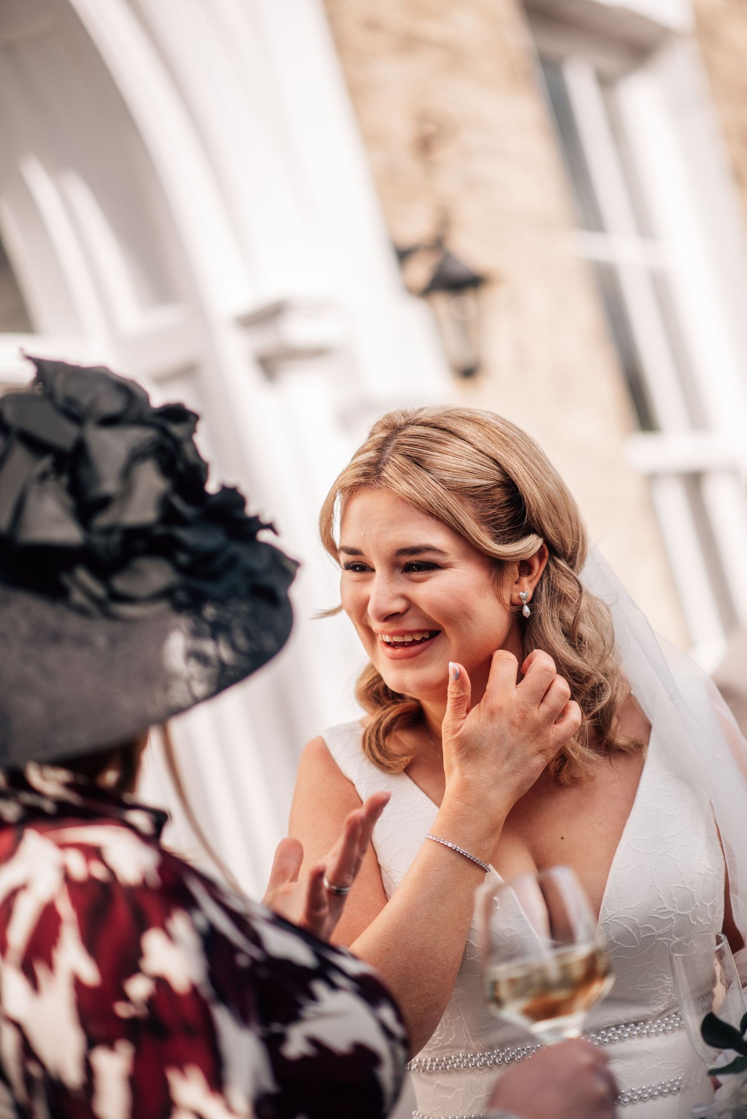 Smiling bride in white dress and veil laughing with a guest in a black hat at an outdoor wedding reception.