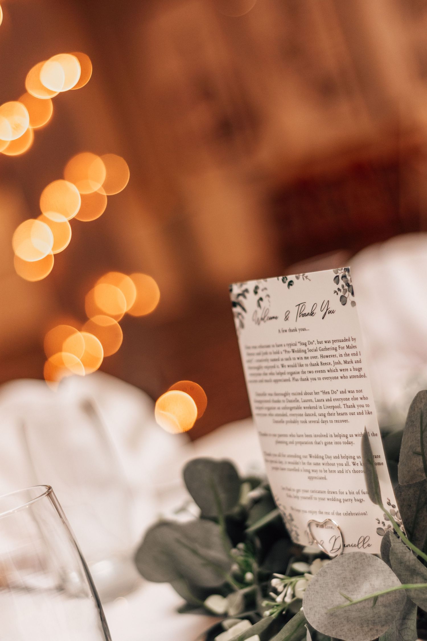 Elegant wedding reception table with a thank you card, eucalyptus greenery, and warm bokeh string lights in the background.