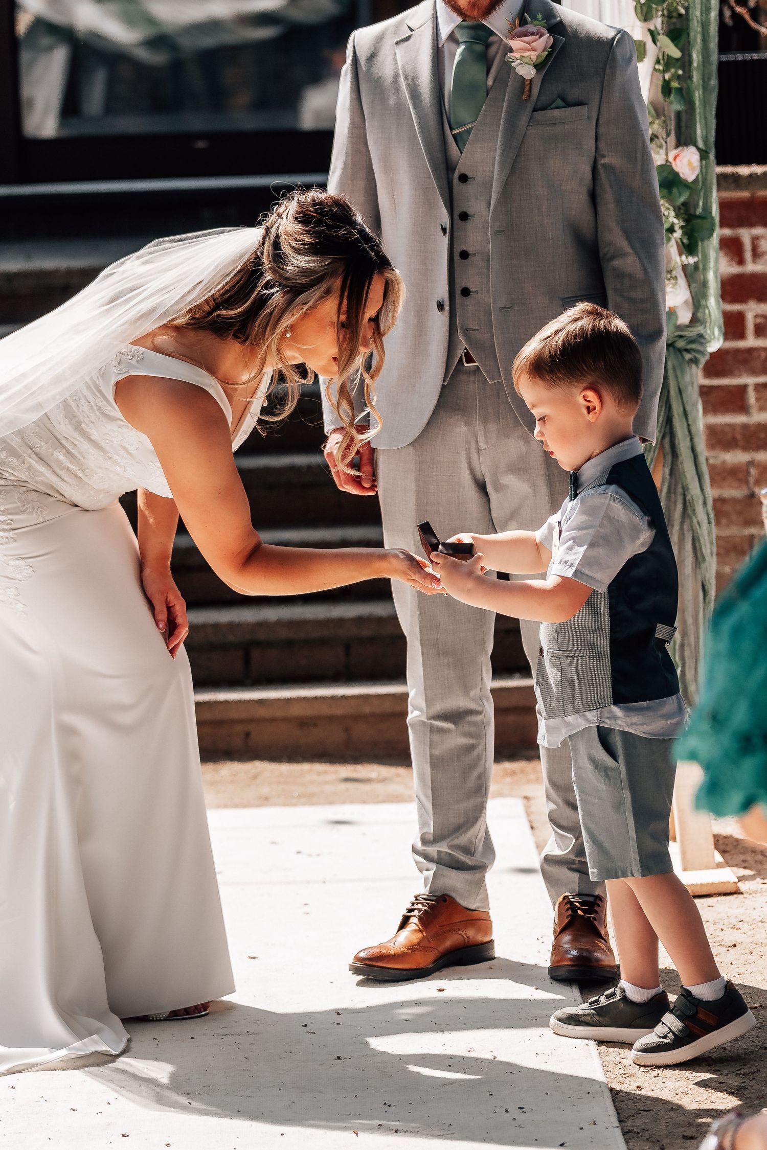 Bride bending down as young ring bearer presents wedding rings during outdoor ceremony with groom standing behind.