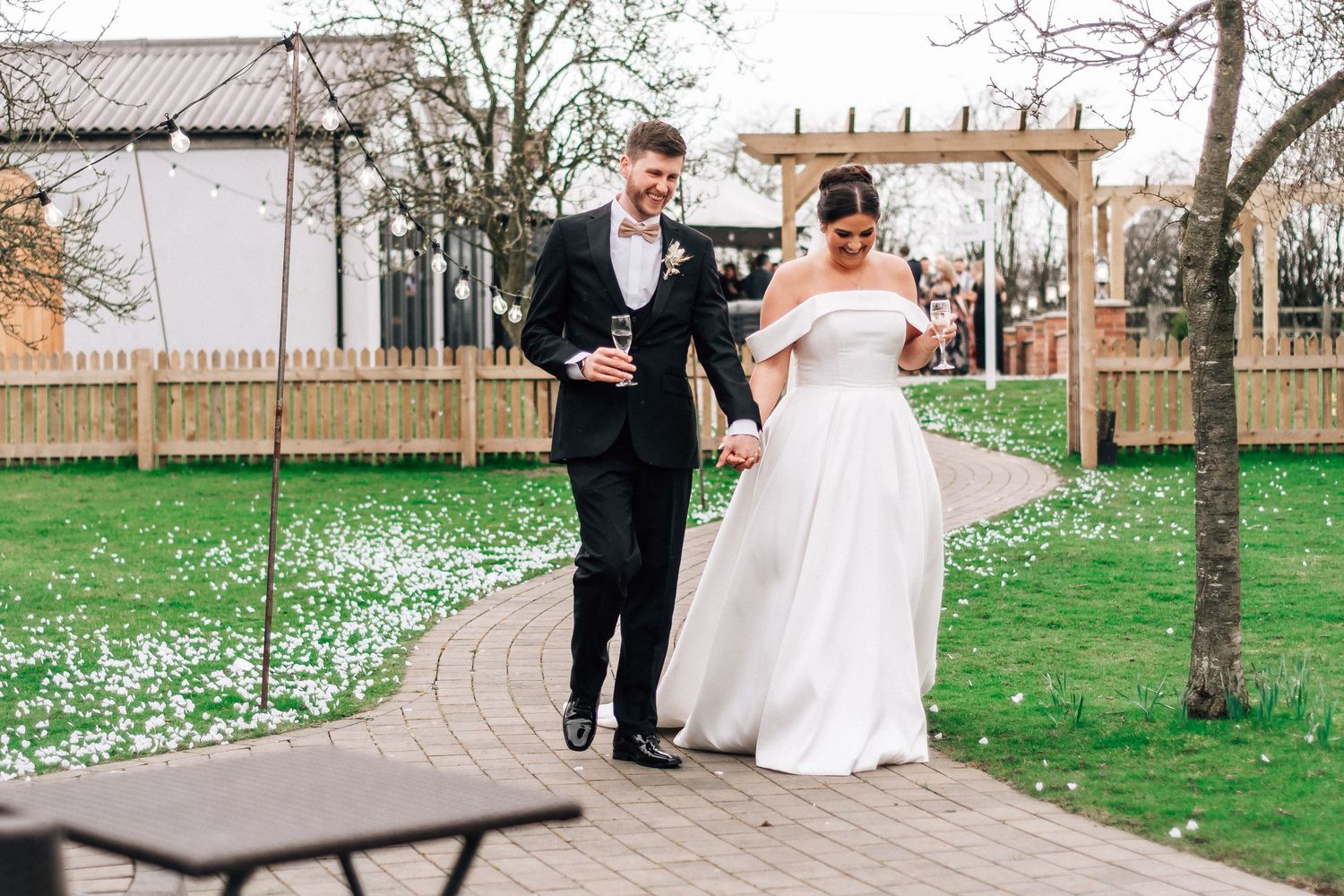 A bride and groom walk hand in hand along a petal-lined path in a lush garden on their wedding day.
