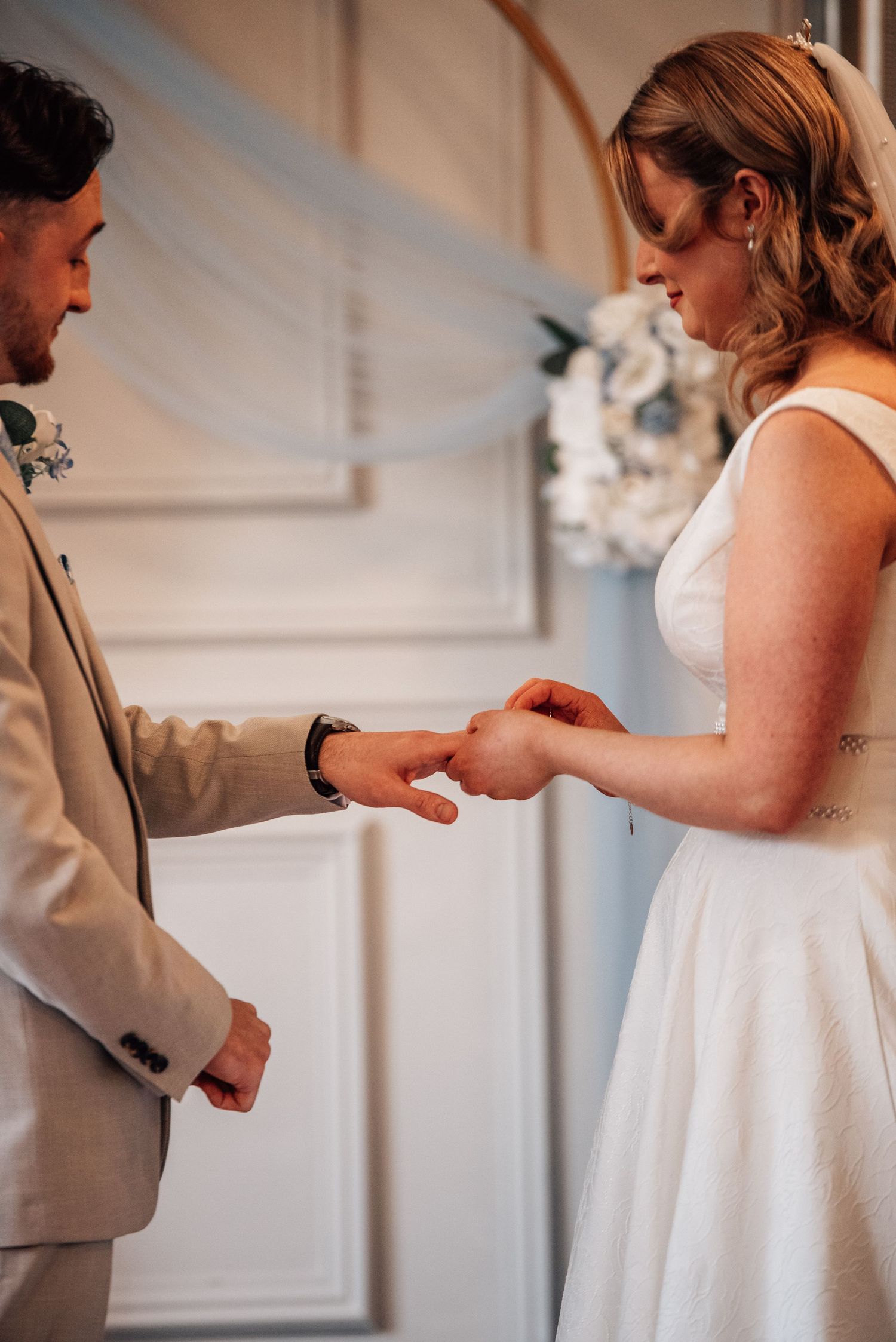 Bride placing a wedding ring on groom's finger during an elegant indoor ceremony, both dressed in wedding attire.