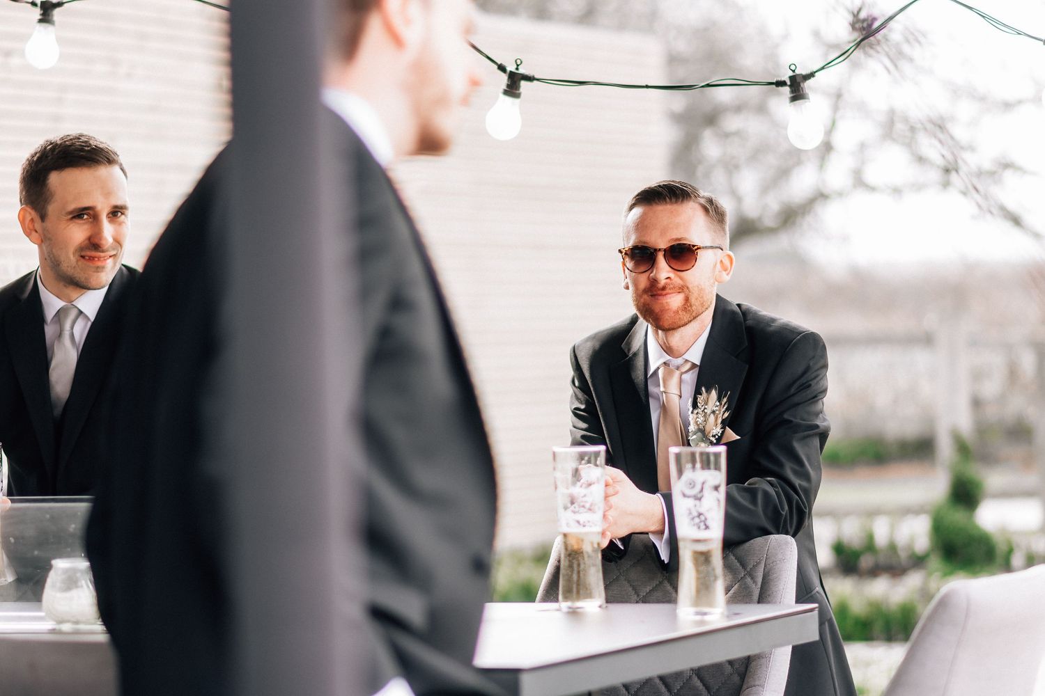 Men in suits socializing at an outdoor wedding reception table with string lights.