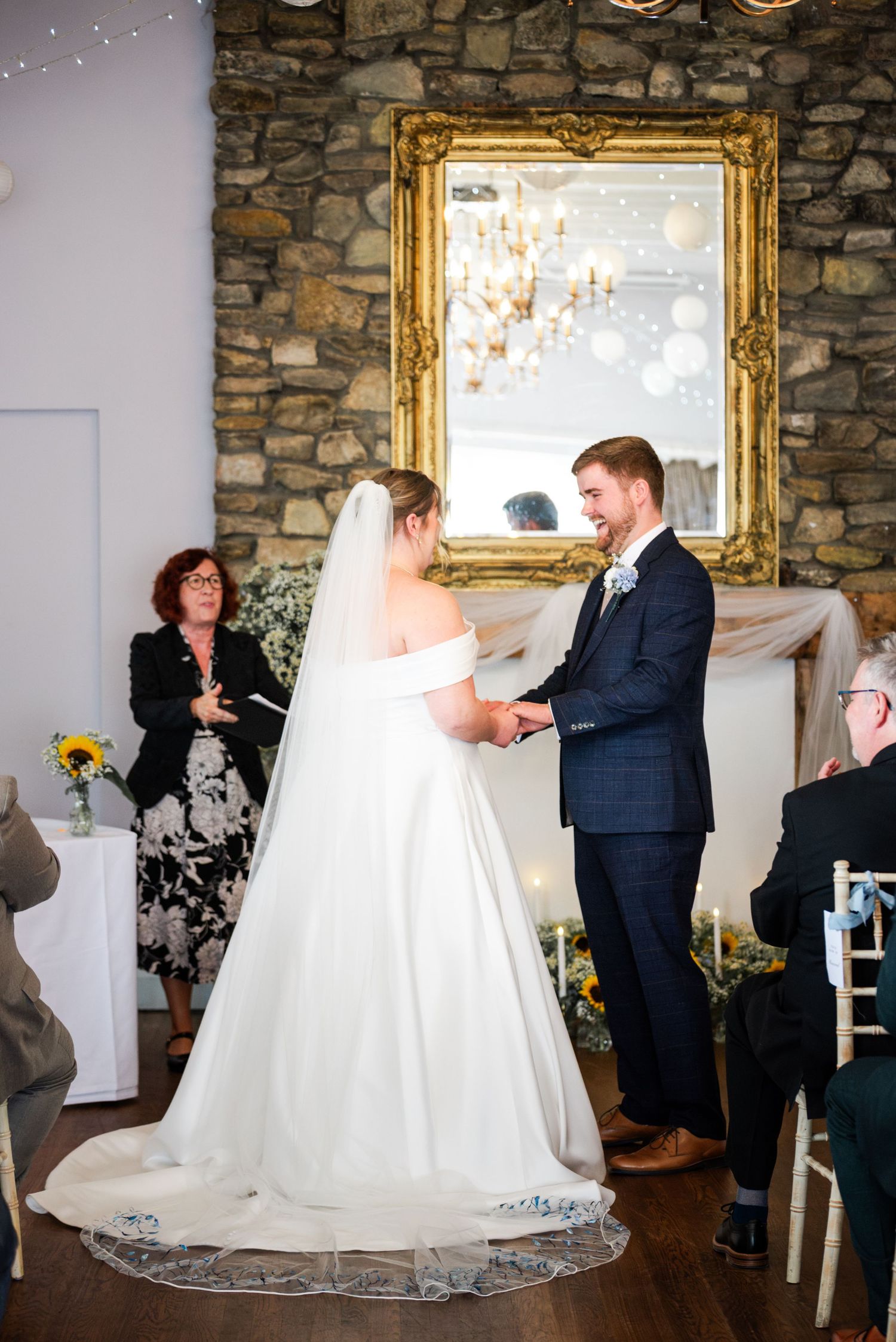 Bride and groom exchange vows at indoor wedding ceremony, stone wall backdrop with ornate gold mirror.