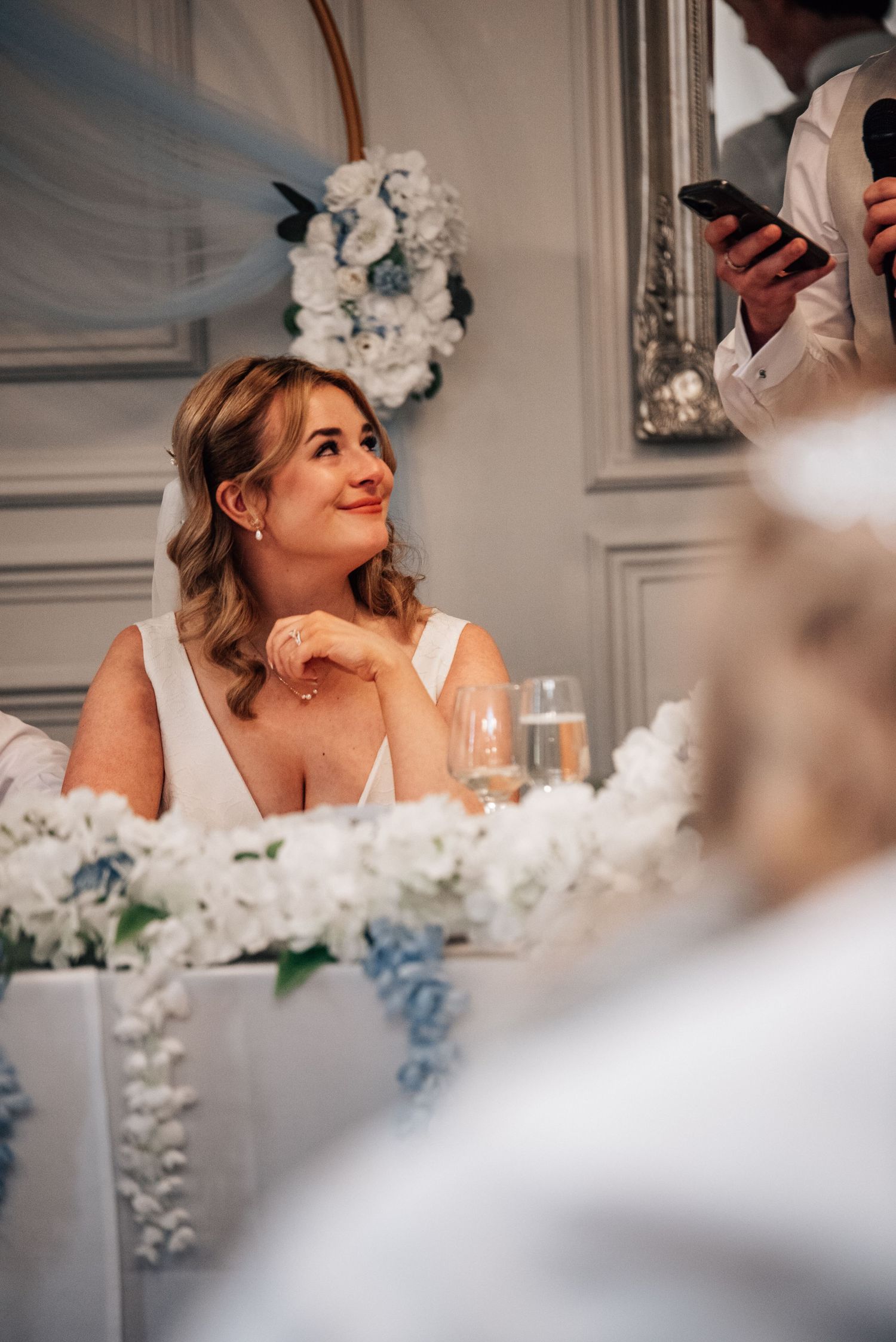 Smiling bride in white gown sits at floral-decorated head table during wedding reception speeches.