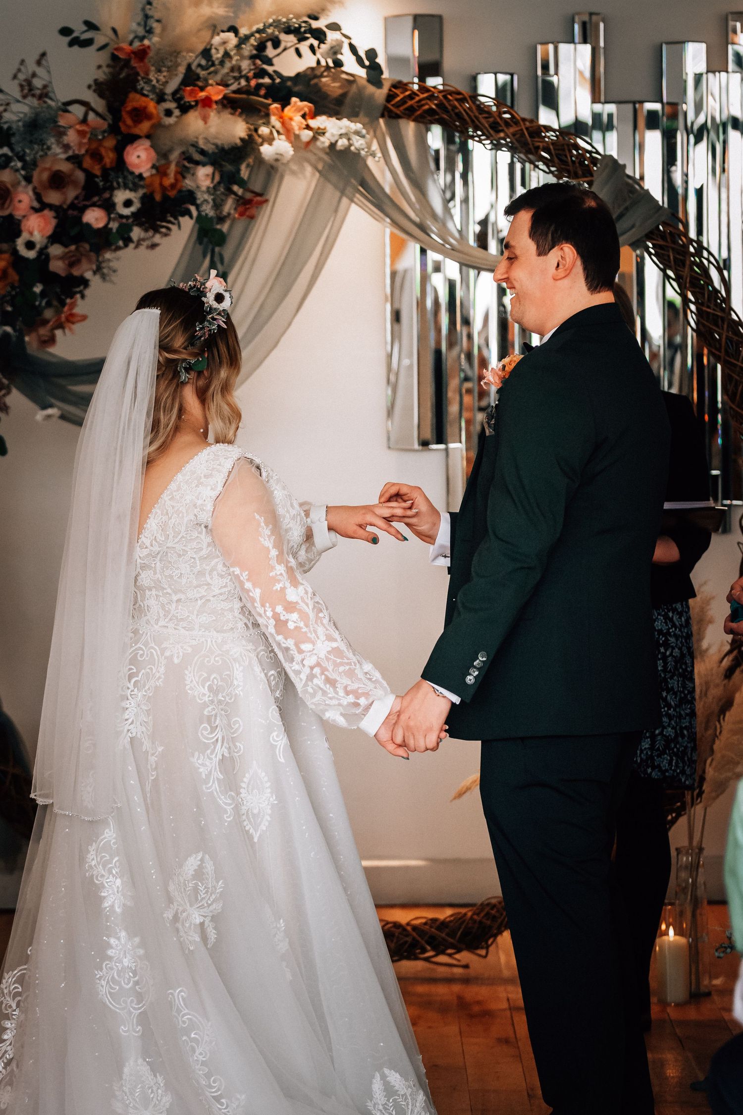 Bride and groom exchange rings at indoor wedding ceremony beneath floral arch with blue and orange blooms.