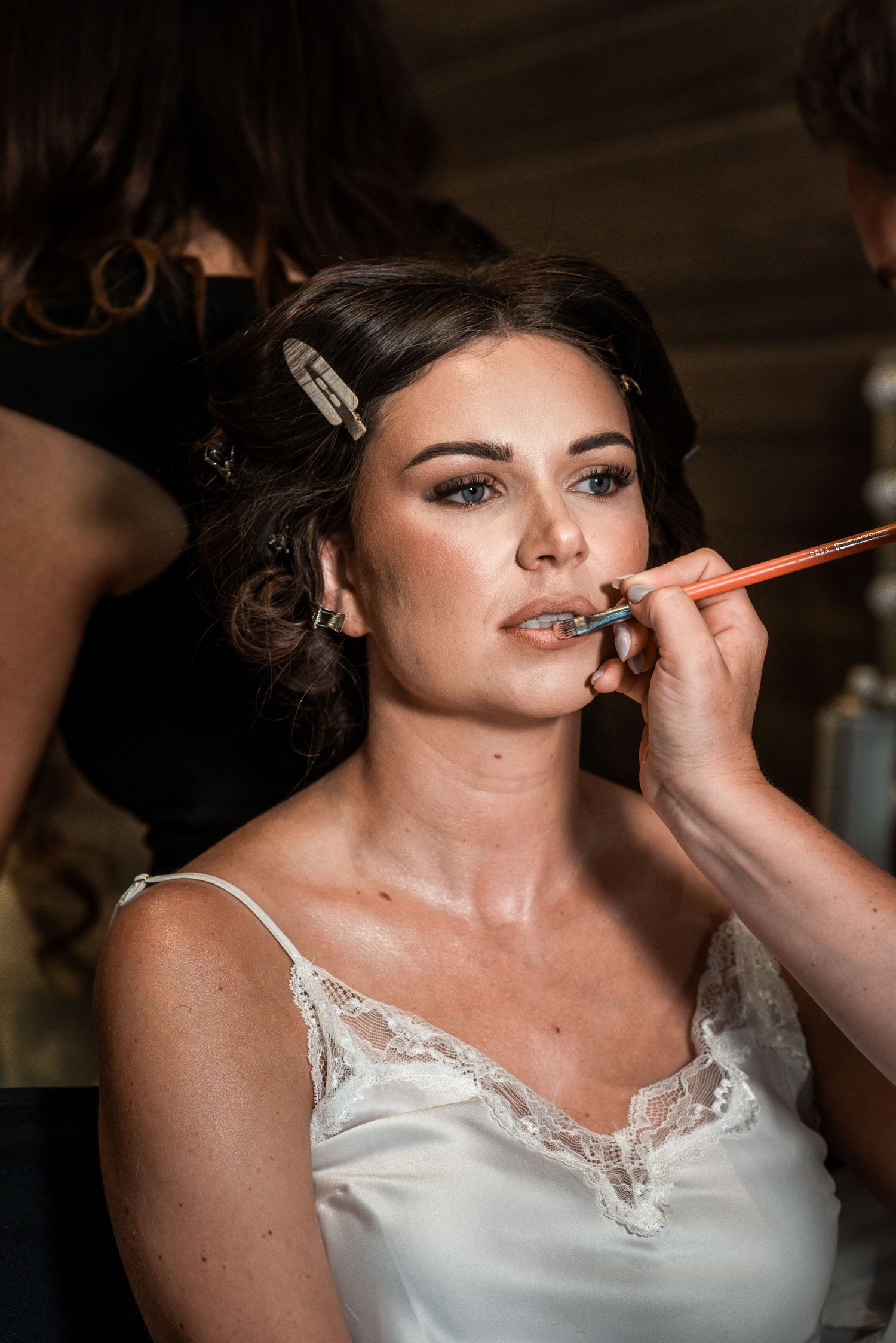 Bride getting makeup applied with a brush while wearing a white lace slip dress before her wedding.