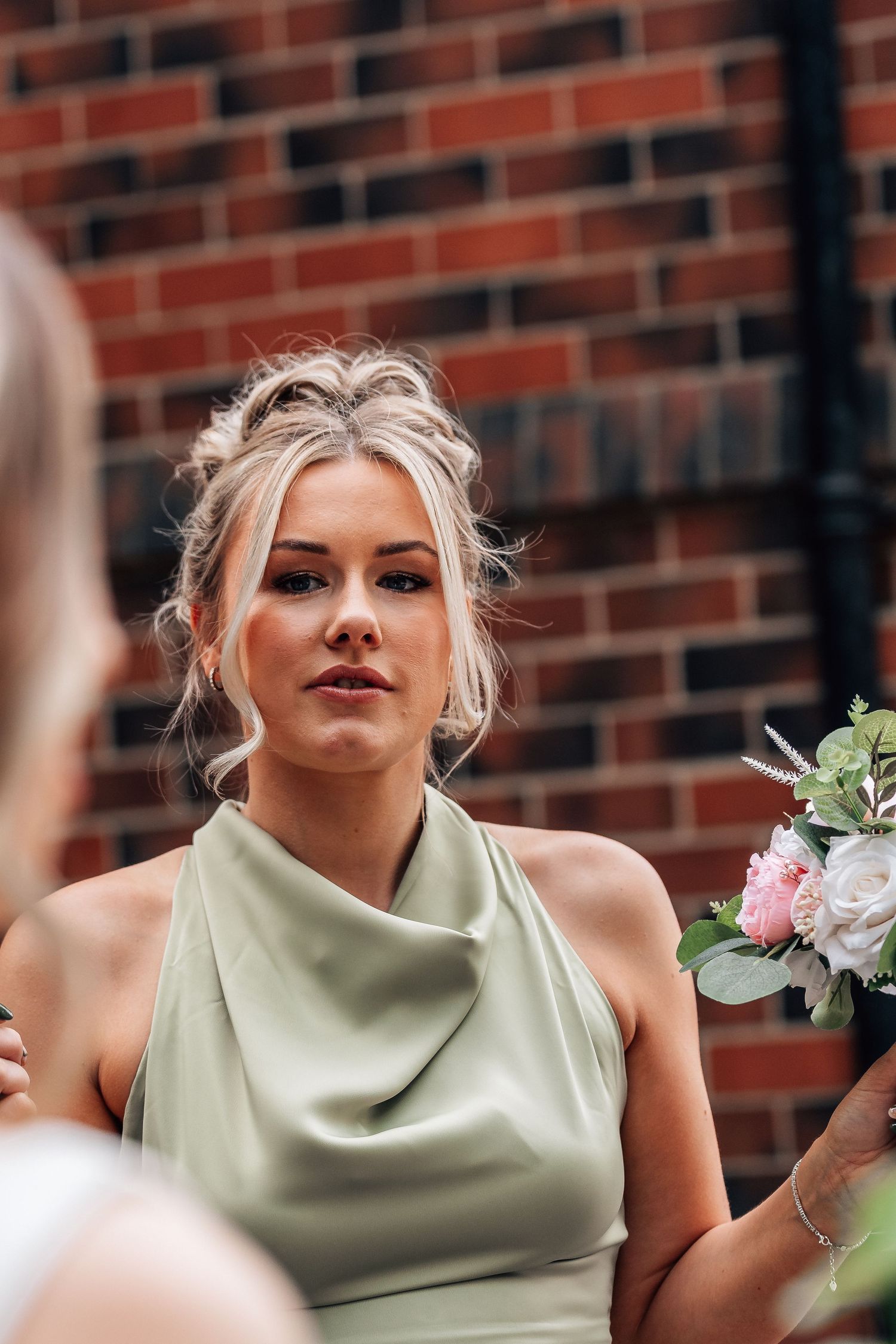 Blonde bridesmaid in sage green satin dress holds pink floral bouquet against red brick wall backdrop.