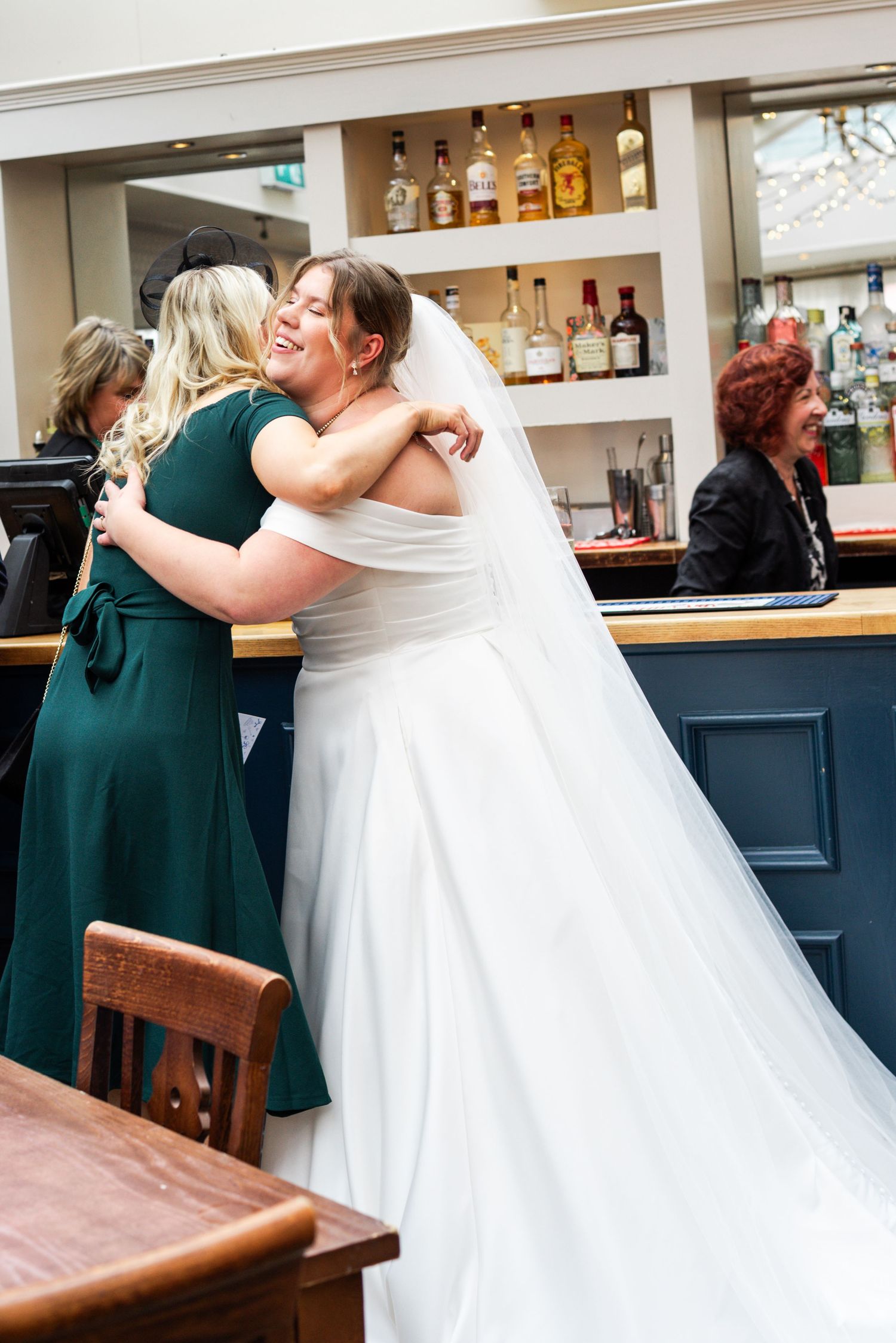 Bride in white gown and veil hugging bridesmaid in green dress near a bar, both smiling joyfully.