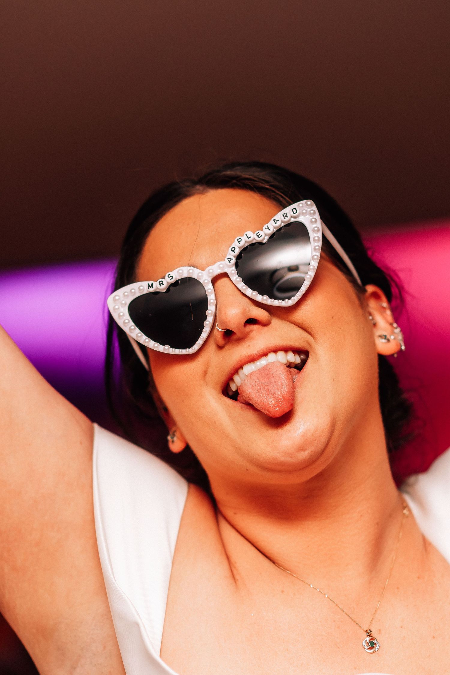 Woman in heart-shaped rhinestone sunglasses sticking out tongue, wearing white top, colorful background.