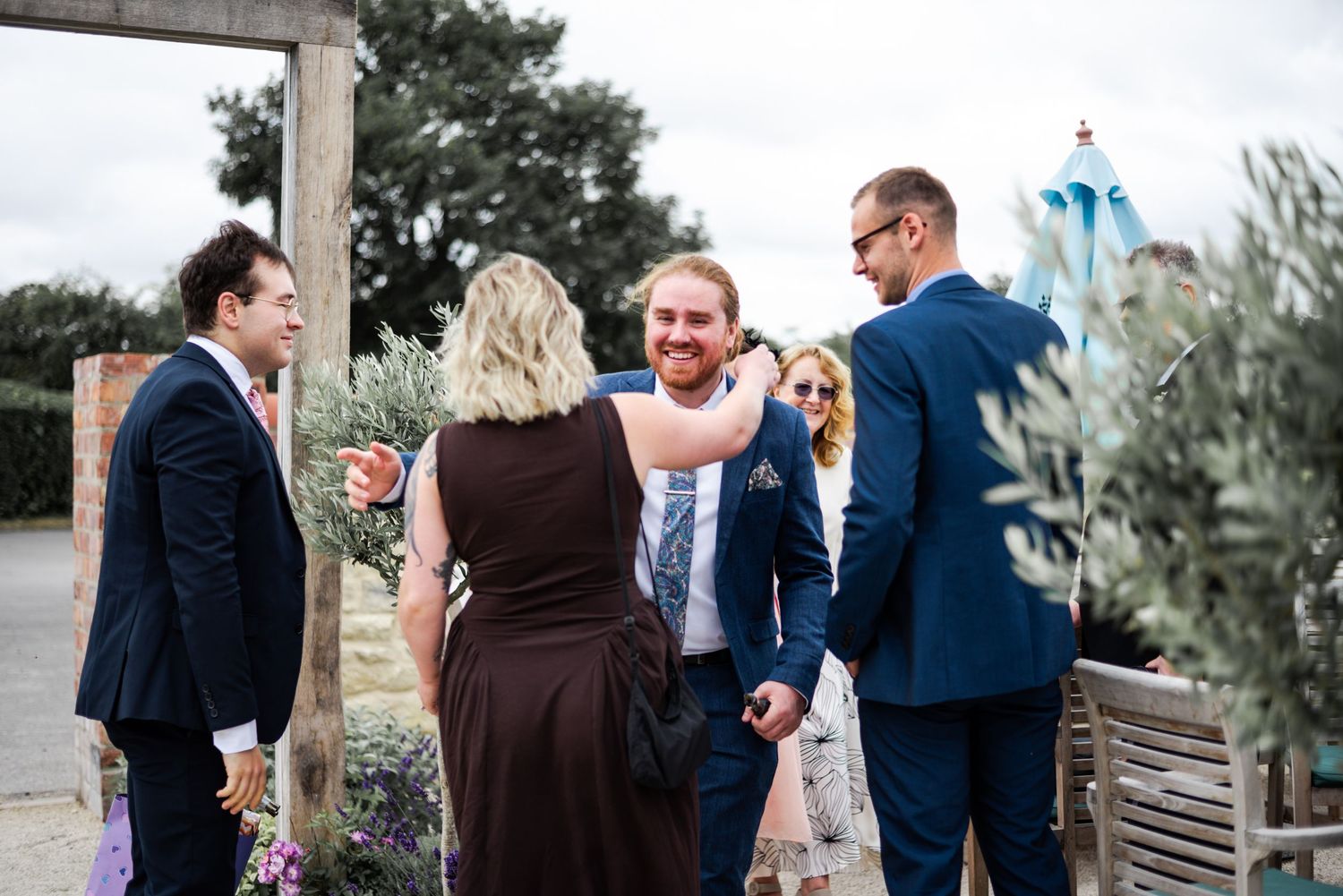 Guests mingling outdoors at a wedding reception, with men in navy suits and a woman in a burgundy dress.