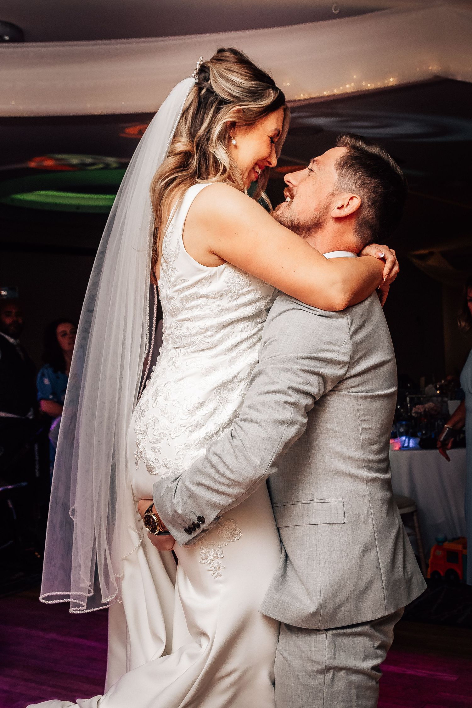 Groom lifts smiling bride in white gown and veil during joyful first dance at wedding reception.