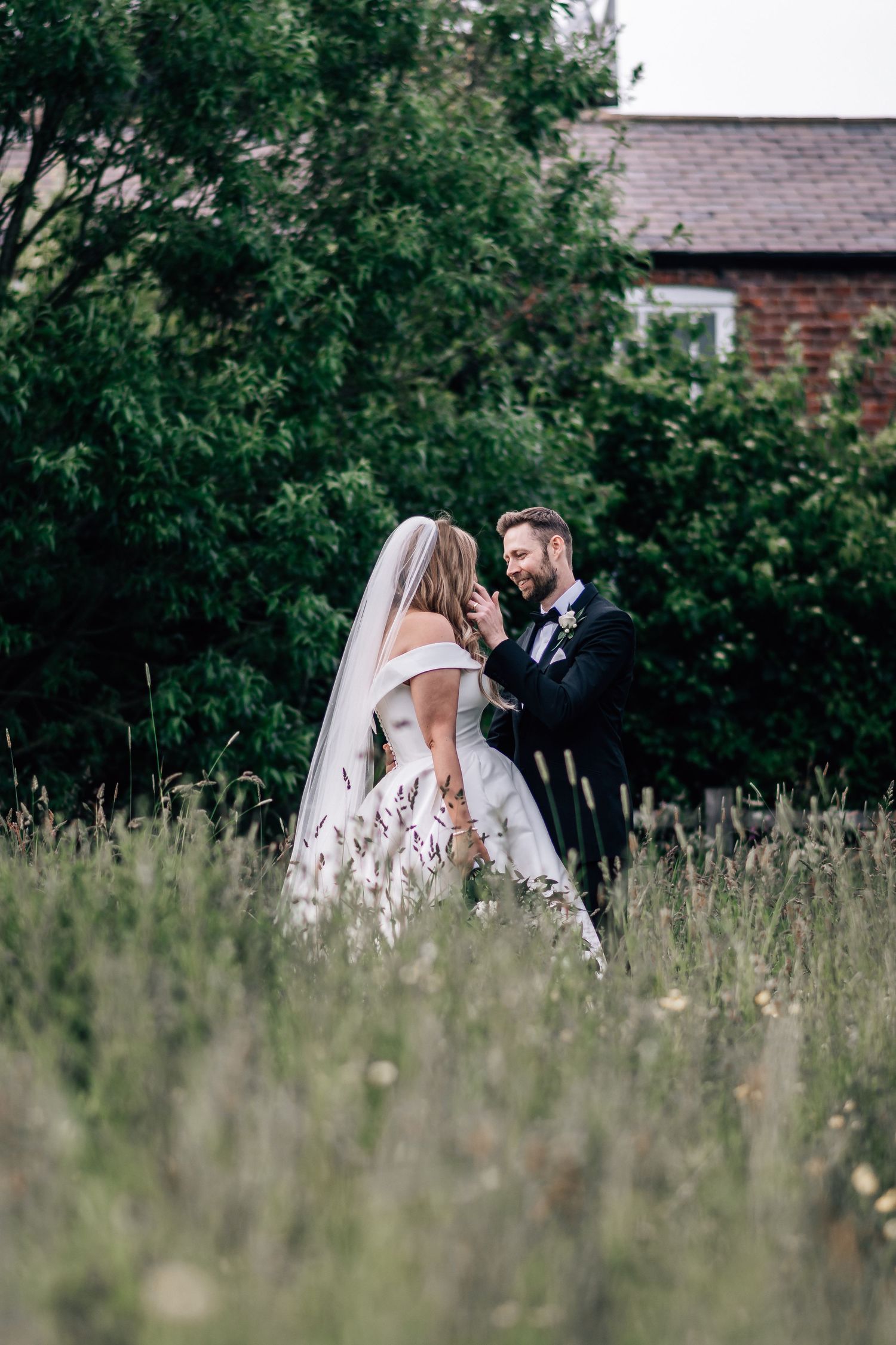 Bride and groom share a romantic moment in a lush green meadow, with the groom tenderly touching the bride's veil.
