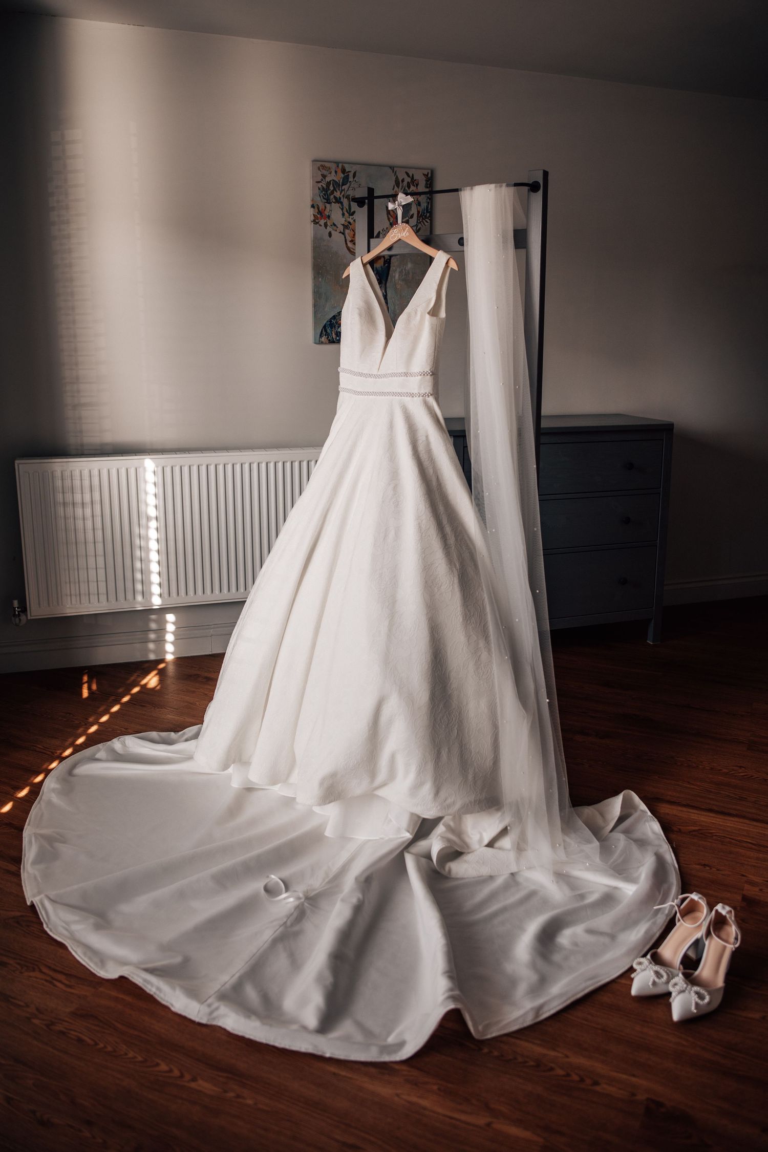 Elegant white A-line wedding dress with deep V-neckline and long train displayed on hanger beside bridal heels.