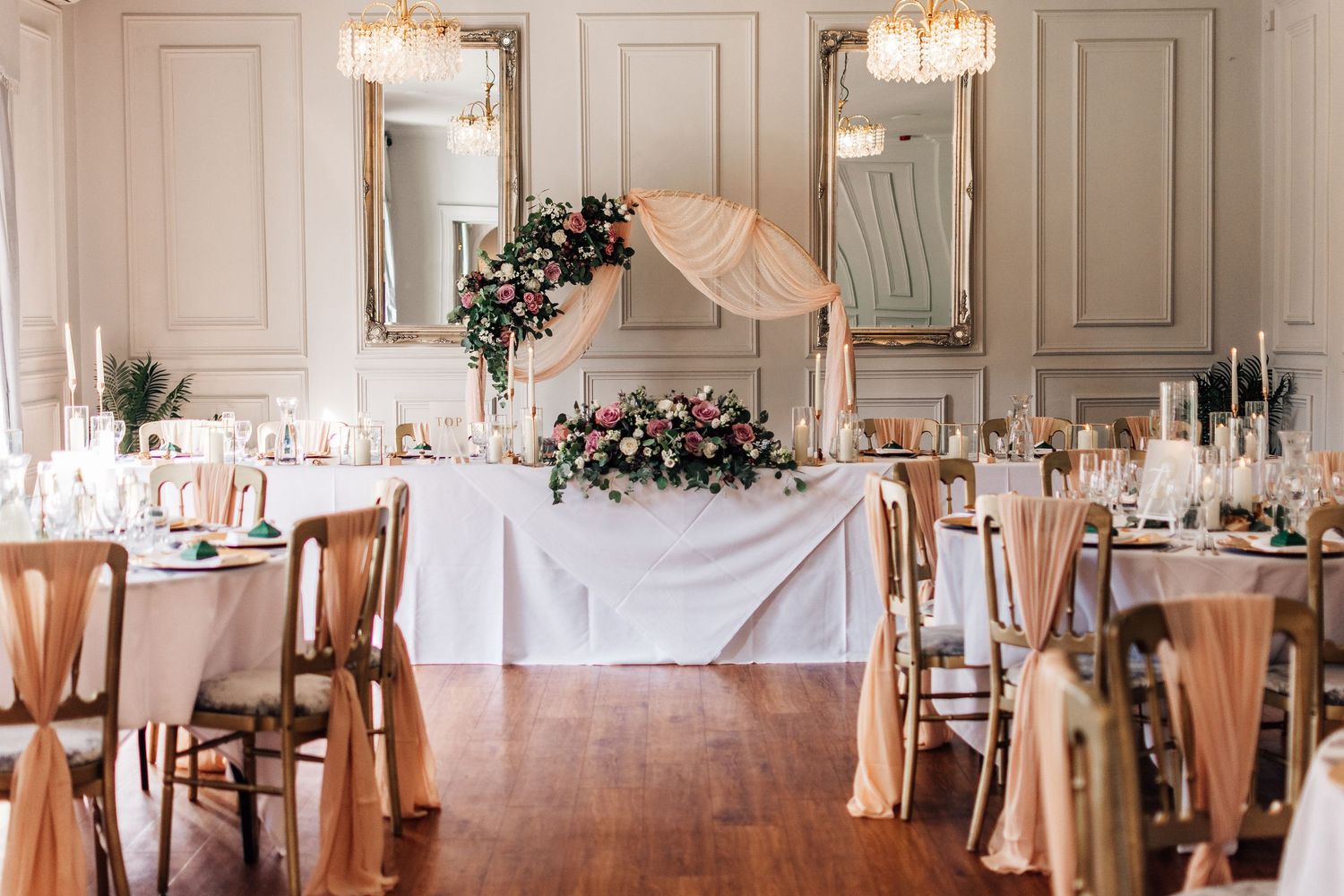 Elegant wedding reception hall with blush pink chair sashes, floral arch, gold mirrors, and candlelit tables on hardwood floors.