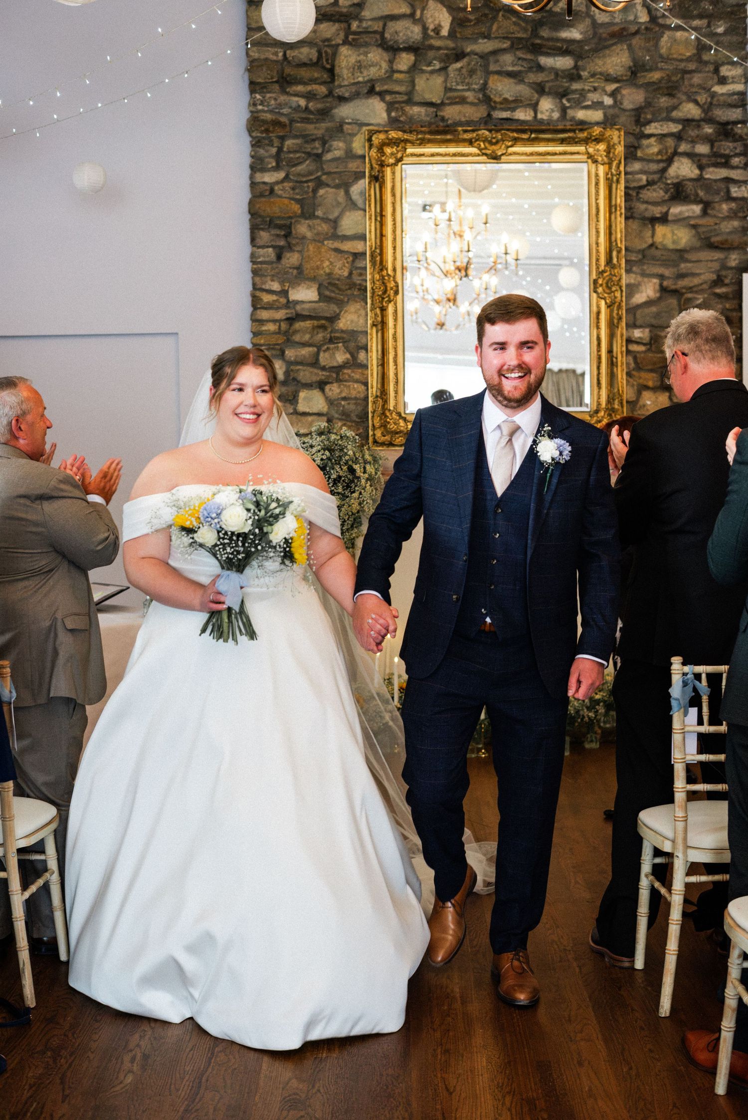 Smiling bride and groom walk down the aisle hand in hand after their wedding ceremony in a rustic stone venue.