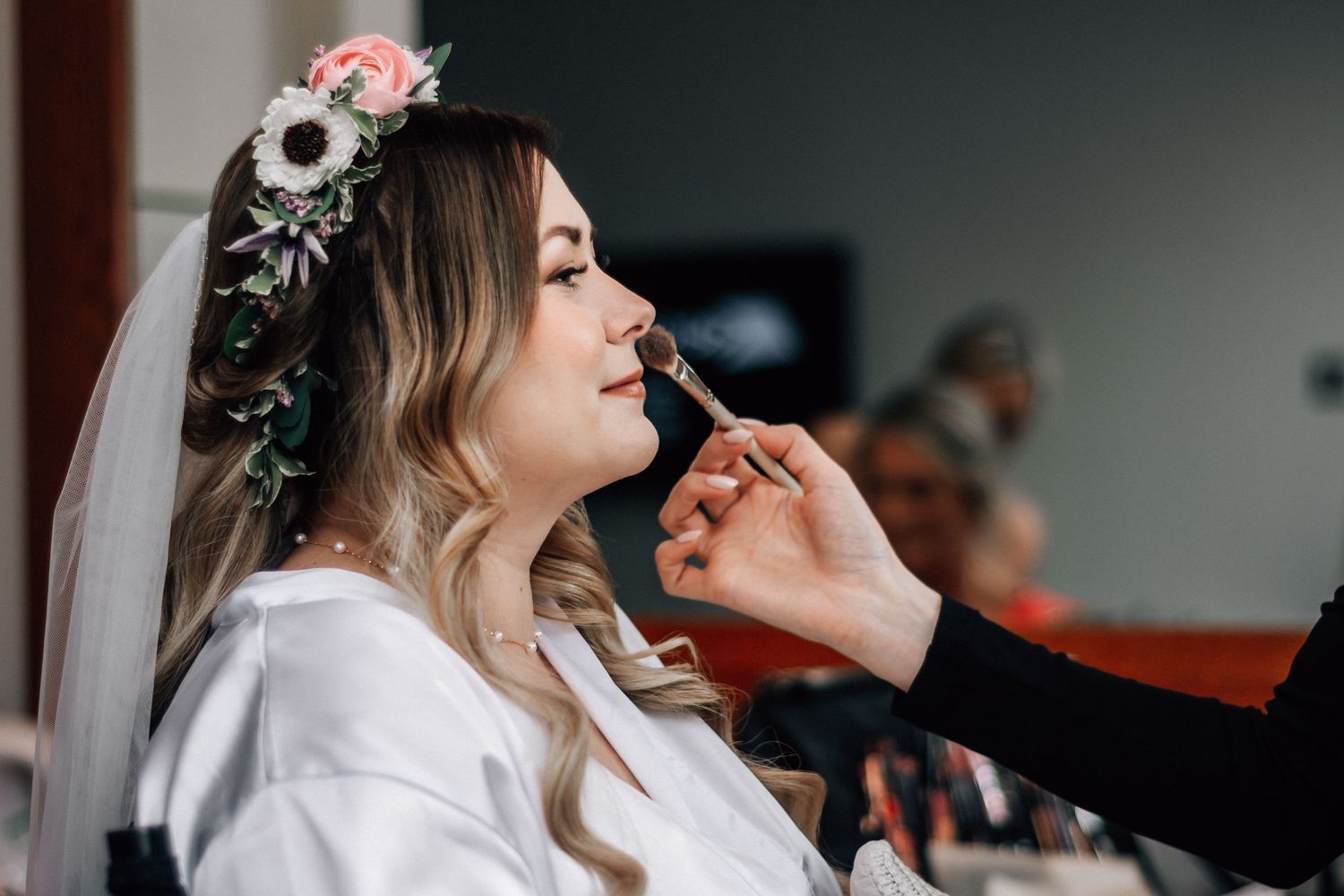 Bride getting makeup applied while wearing floral crown and veil on wedding day.