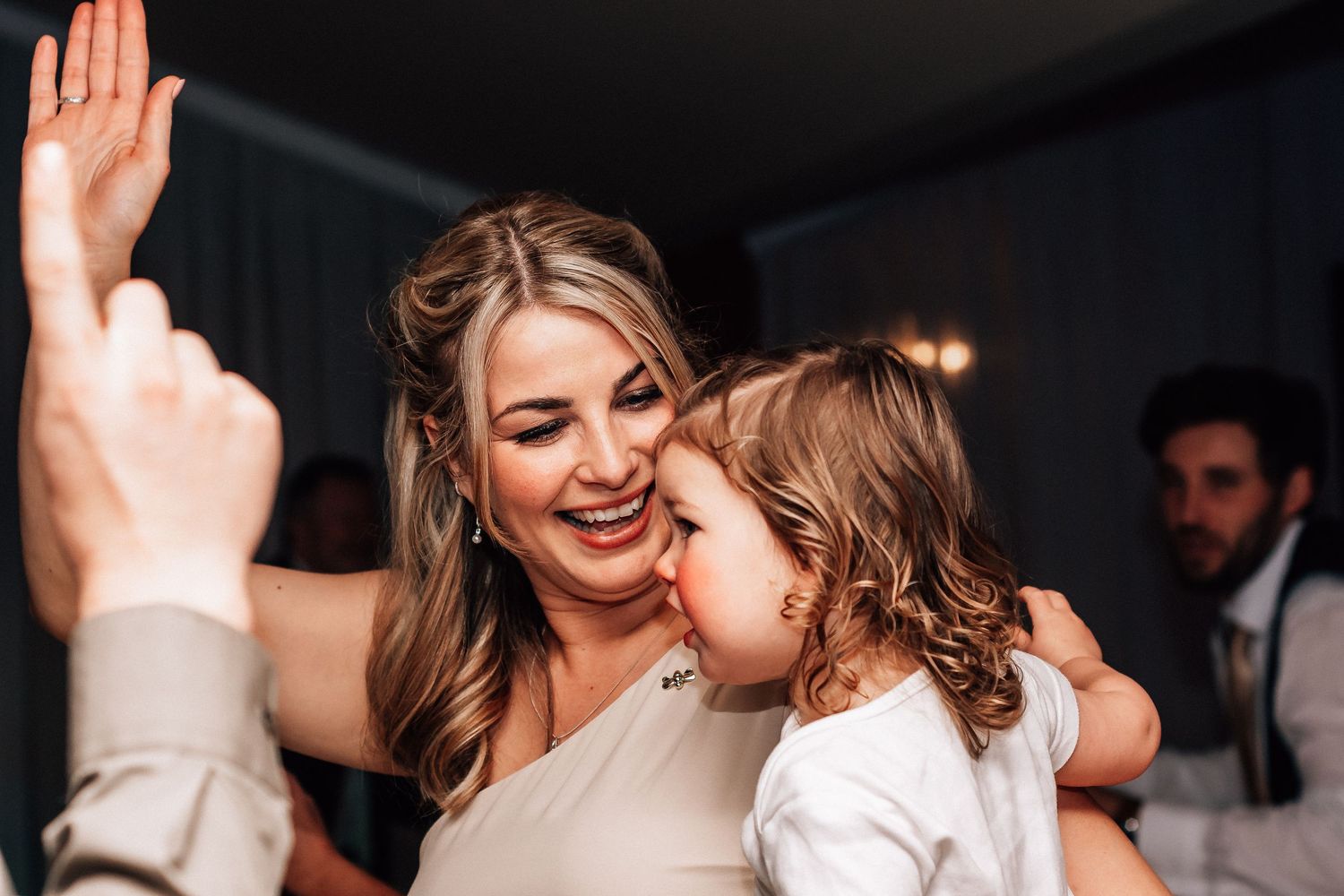 Smiling bride in white dress holding a curly-haired toddler at a wedding reception dance floor.